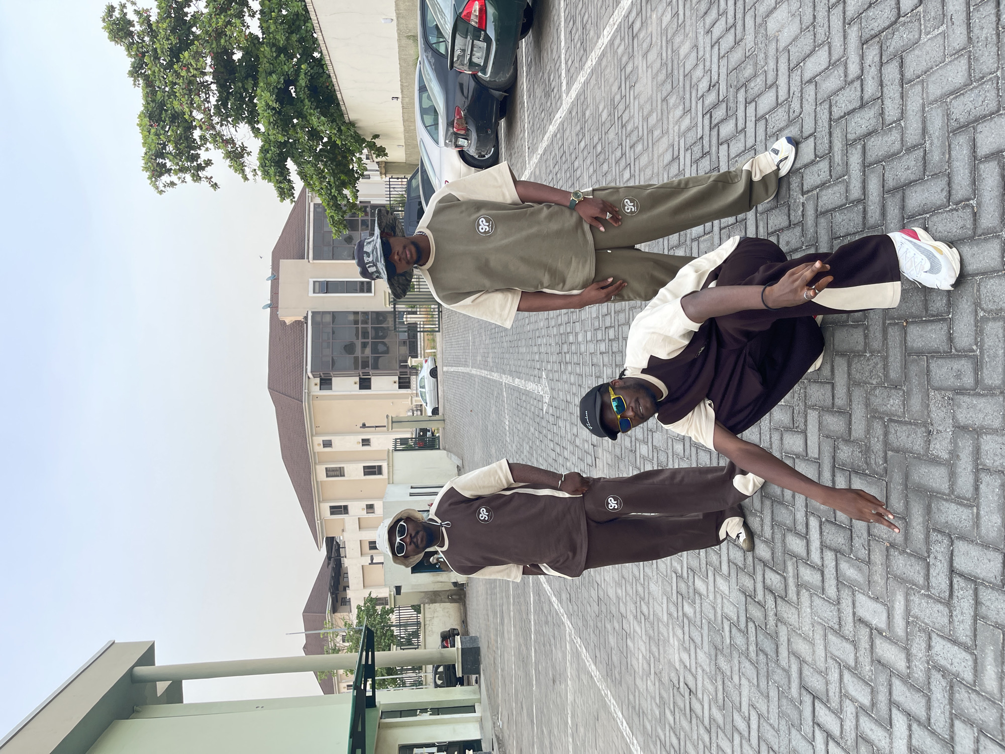 The image displays three individuals, likely young men, posing in an outdoor, urban setting in Abuja, Nigeria. The scene is a paved area, possibly a parking lot or courtyard, with modern buildings, parked cars, and green trees in the background. The clear, bright sky suggests it is daytime.
Two of the individuals are standing upright, while one is in a crouching pose. They are all wearing coordinated casual outfits consisting of a t-shirt and loose-fitting pants. One person wears an olive green set with cream-colored accents, while the other two wear brown sets, also with cream accents. A distinctive circular logo with the letters "SP" is visible on the chest of their shirts and on the left thigh of their pants.
Accessories vary among the individuals: one wears a patterned bucket hat and a wristwatch; another has a black baseball cap (with some unreadable text) and sunglasses perched on it; and the third wears a light-colored bucket hat and white-framed sunglasses. All subjects are wearing sneakers, with visible branding on some, including a Nike-like swoosh on one pair.
The ground is covered with grey interlocking pavers laid in a herringbone pattern. In the background, multi-story buildings with light-colored facades and dark roofs are visible, alongside parked vehicles, including a dark sedan. Lush green tree foliage extends into the upper left of the frame. The individuals appear to be confidently posing for the camera.