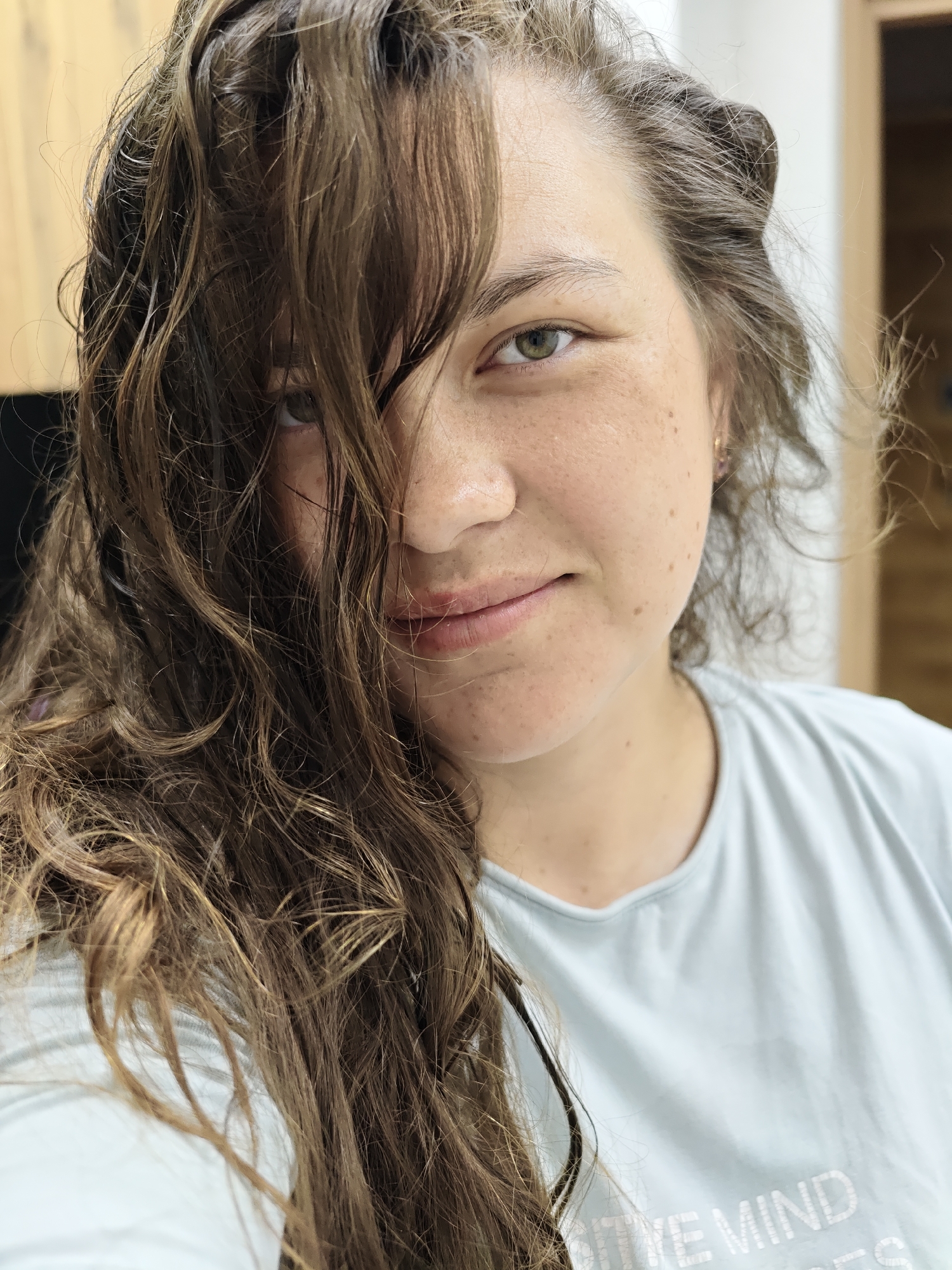 A selfie portrait of a fair-skinned woman with brown hair partially obscuring her face. Her hair is wavy and appears to be slightly damp. She has freckles and green eyes. She is wearing a light blue t-shirt with the words "POSITIVE MIND" visible. The background is out of focus, suggesting an interior setting. The location is not indicated and cannot be determined from the visual information.
