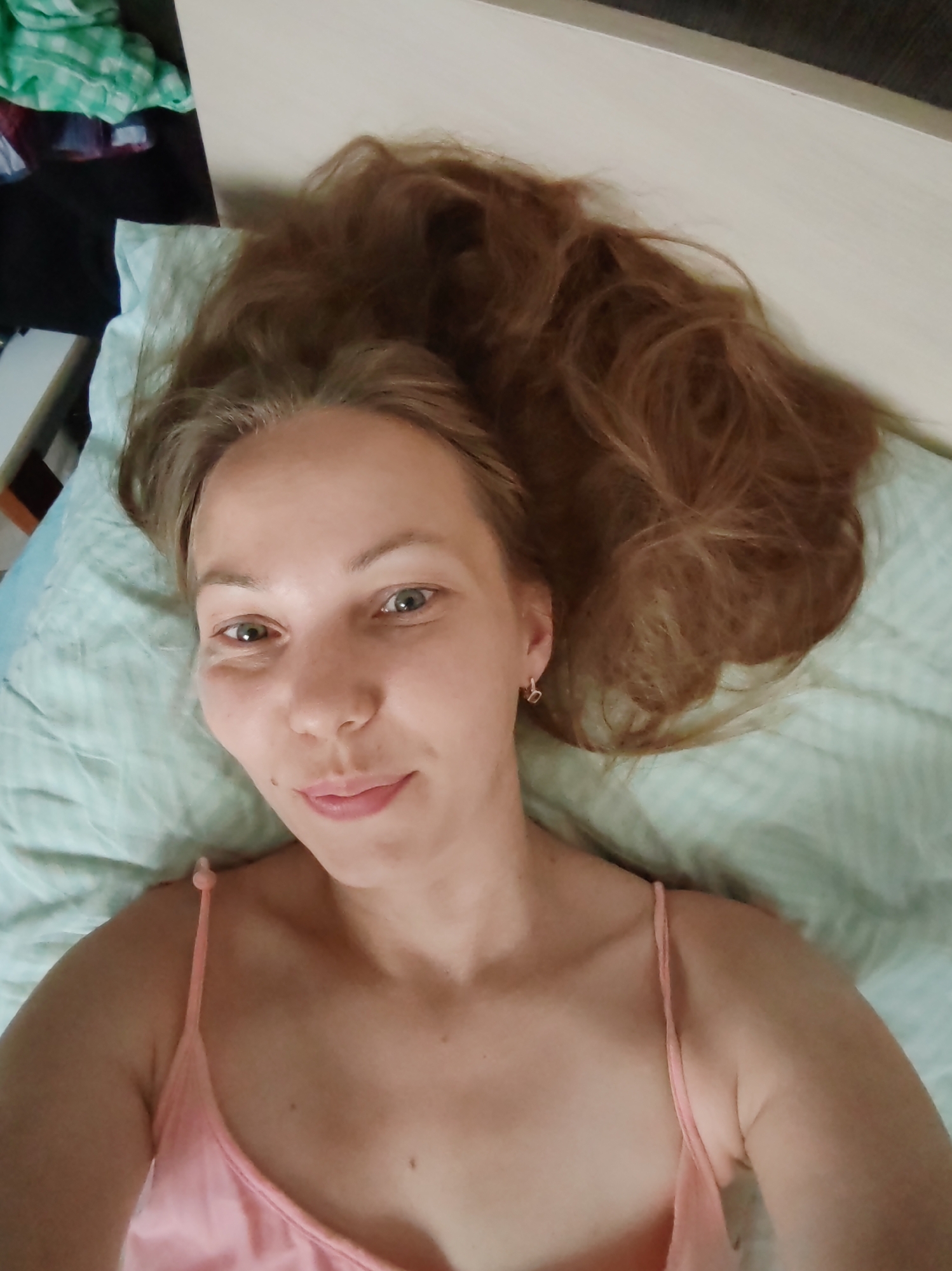 A young woman with long, light brown hair lies on her back on a light blue and white striped bed. She is wearing a pink spaghetti strap top. The woman's face is turned towards the camera, and she is smiling slightly. Her eyes are open and looking at the camera. Her right eye appears to have some redness or swelling around it. She has a small mole on her left cheek. She is wearing a small silver hoop earring in her left ear. In the background, to the left, are piles of colorful clothing and a white dresser or shelf. To the right, the bed extends into the background. The lighting suggests it is daytime, and the setting is likely a bedroom. The location is specified as Чухлэм, Russia, though this cannot be visually confirmed from the image itself.