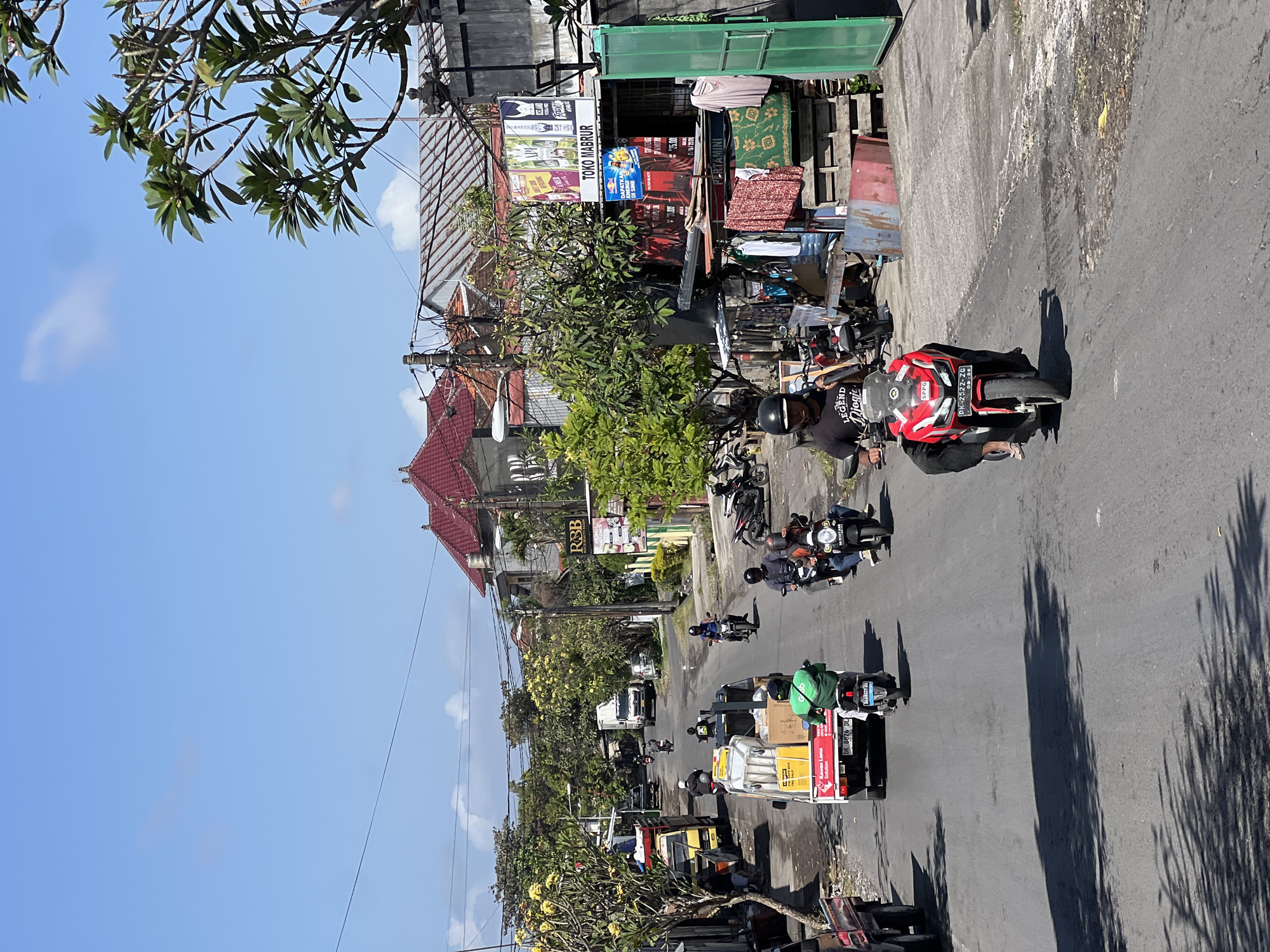 This is a medium-angle, eye-level, full shot of a street scene in Denpasar, Indonesia, on a sunny day. The street is paved, with a slight incline and a gutter running along the right side. On the left, there is a parked truck carrying boxes and other items, with a person standing next to it. Further down the street on the left, more trucks and motorcycles are visible. The right side of the street is lined with buildings, including a shop with clothing racks displaying garments and a sign that reads "TOKO MABRUR". Various motorcycles are parked and being ridden on the street. Several individuals are seen riding motorcycles, some wearing helmets. Trees with green foliage and yellow flowers are present on both sides of the street, and buildings with red tiled roofs are visible in the background. The sky is blue with scattered clouds.