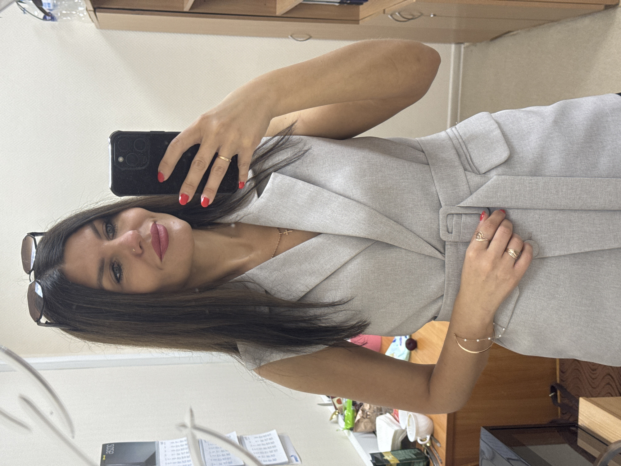 A woman with long, dark hair and sunglasses perched on her head takes a mirror selfie. She is wearing a light gray sleeveless blazer with a belt and is holding a black smartphone in her right hand. She has red nail polish on her fingernails and is wearing rings on her fingers. A delicate gold necklace with a cross pendant is visible around her neck, and a thin bracelet adorns her left wrist. The background shows an office environment with a desk, shelves, and calendars on the wall. The scene is indoors, likely in an office in Chaikovsky, Russia.