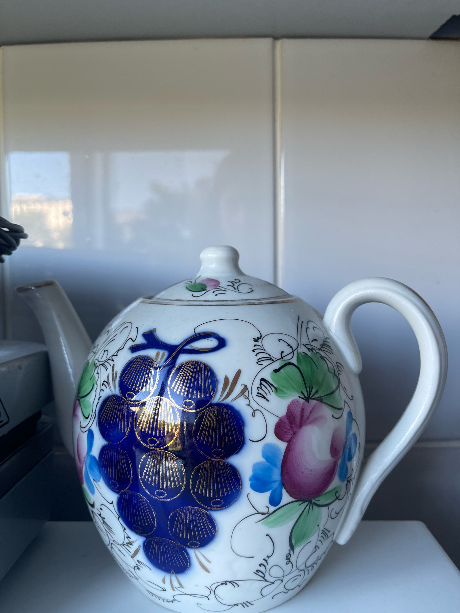 A white porcelain teapot with a grape and floral design is positioned on a white shelf against a background of white tiles. The teapot features clusters of dark blue grapes with gold detailing and pink and blue flowers with green leaves. The handle and spout of the teapot are white. The background tiles reflect light and show a blurred outdoor scene. The image is cropped to focus on the teapot. The teapot is located in Tallinn, Estonia.