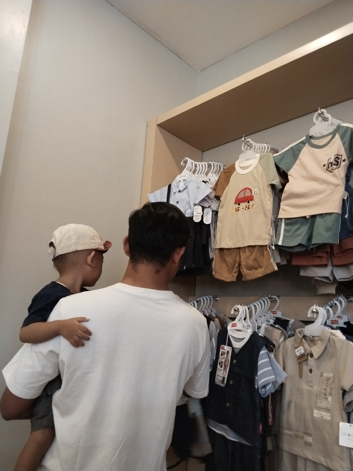 A man with dark hair, wearing a white t-shirt, carries a young boy with a light-colored baseball cap on his left hip in a retail store. The boy, wearing a dark blue t-shirt, looks to his left at the clothes displayed on shelves. The shelves are filled with children's clothing on hangers. The clothing consists of shirts, shorts, and coordinated outfits. The background is a plain, light-colored wall. The lighting is bright, coming from overhead fixtures. The store appears to be a clothing boutique. The scene is likely in Kecamatan Pati, Indonesia, given the provided context.
