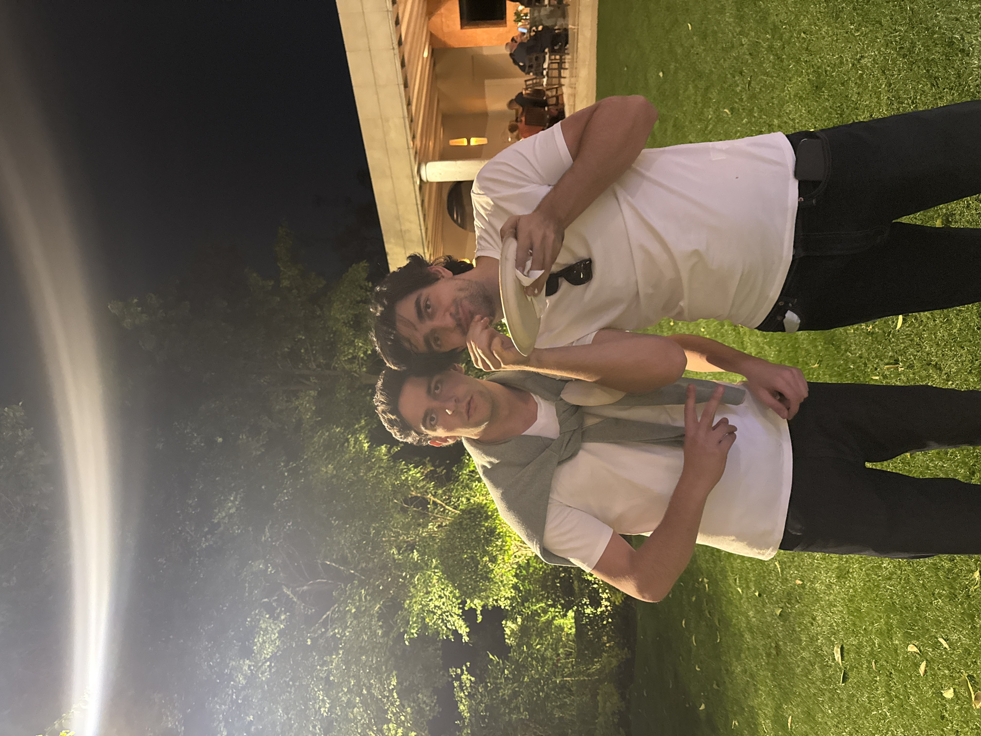 Two men are standing outdoors at night, likely posing for a photograph. The location appears to be a lawn area next to a building with a colonnade. One man is holding a plate and sunglasses, while the other is gesturing with a "peace" sign. The background suggests an outdoor dining area. The location is potentially in Mexico City, Mexico.