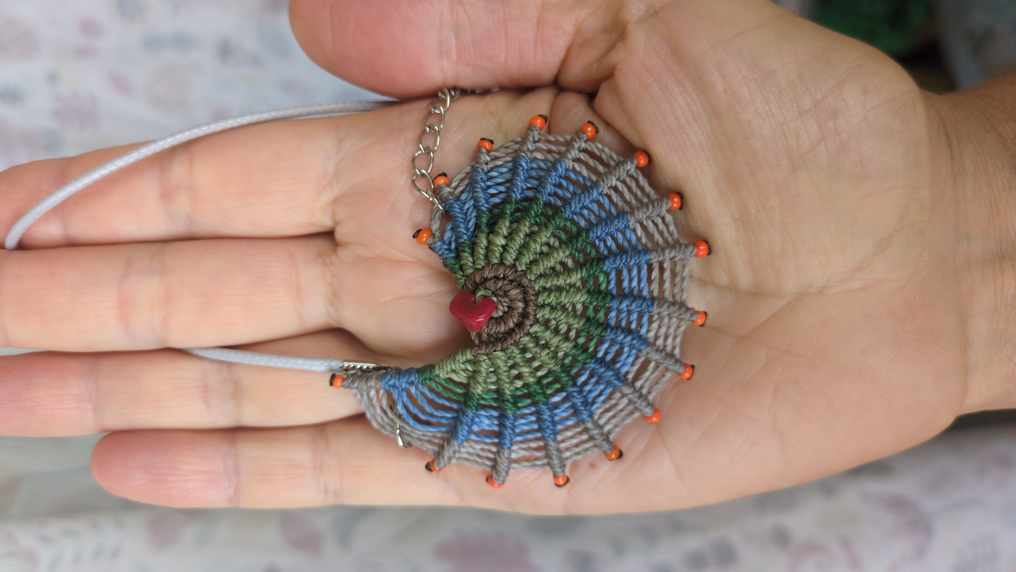 A close-up shot shows a person's left hand holding a macrame pendant. The pendant is fan-shaped, with alternating bands of grey, blue, and green thread. It features an orange bead at the tip of each radiating thread and a red heart-shaped bead in the center. A thin, light-colored cord is attached to the top of the pendant, with a delicate silver chain visible on the right side. The hand is resting against a softly blurred background with a floral pattern. The image does not contain any information about the location in New York City, United States of America.
