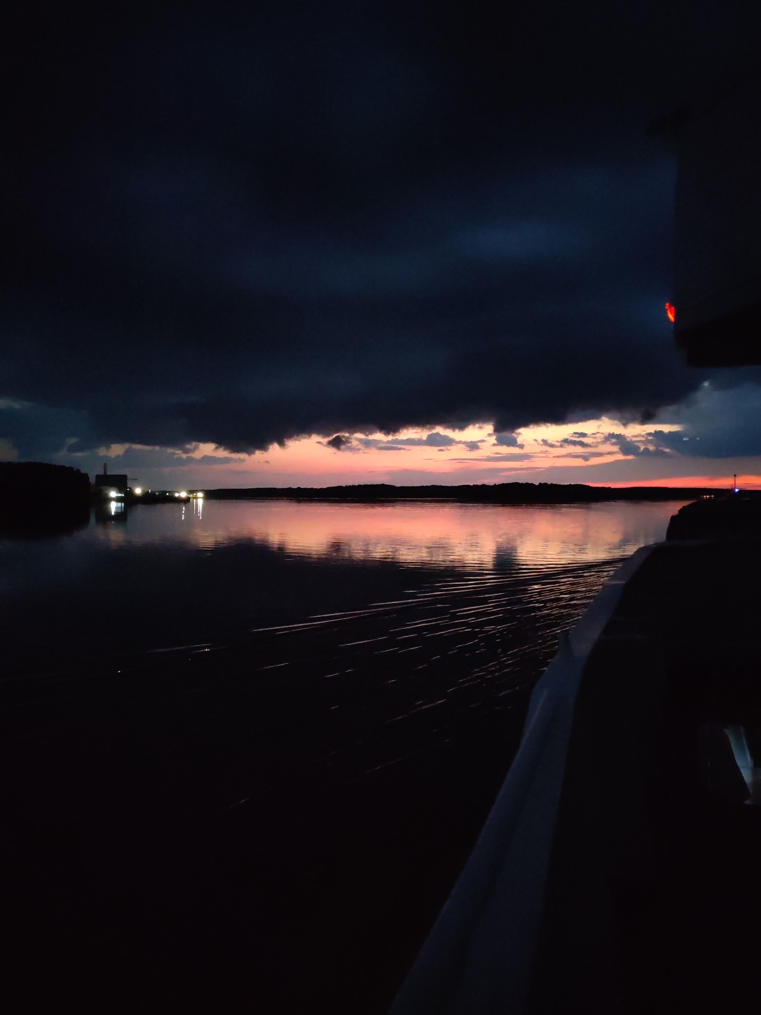 The image depicts a body of water at dusk or dawn, with the sky displaying a gradient of dark clouds transitioning to soft pink and orange hues near the horizon. A silhouetted treeline marks the far shore, punctuated by several bright lights, possibly from buildings or structures. The water reflects the colors of the sky and the artificial lights, creating a shimmering effect. In the foreground, the dark, rippling wake of a vessel is visible, suggesting the image was taken from a moving boat. Part of the vessel's structure, dark and indistinct, occupies the right side of the frame, with a faint red light visible at its upper edge. The overall atmosphere is serene and twilight-like, characteristic of a northern latitude location such as Bol'shie Mkhi, Russia, during a time of low light. There are no people or specific actions readily discernible.