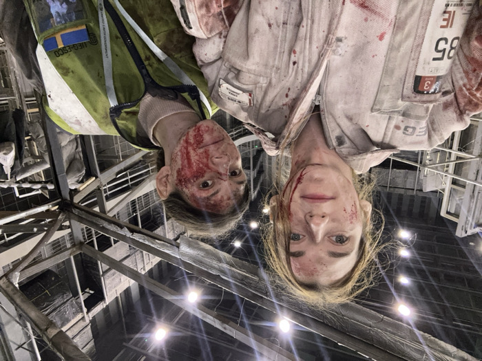 The scene depicts two individuals in an indoor facility or set, identified as being in Huangdao District, China. Both subjects are positioned facing towards the camera.
The individual on the left, partially in the foreground, has light brown hair. This subject is wearing a light-colored, possibly white or light grey, utility jumpsuit, which is heavily stained with red liquid resembling blood, distributed as splatters and streaks across the torso and arms. Red liquid is present on the face, specifically on the left cheek and forehead. Identifiable patches on the jumpsuit include 'V3', '85 IE', and 'EMMA JOHNSON', along with partial text 'EG-'.
The individual on the right, positioned slightly behind and to the right of the first subject, has darker hair. This individual wears a light-colored long-sleeved shirt beneath a yellow-green high-visibility vest. The vest is also marked with red liquid stains. The face is extensively covered with red liquid, concentrated on the right side. A patch on the vest displays 'UFG-ESCO' with a globe emblem and a blue square featuring a yellow cross, consistent with the flag of Sweden.
The background reveals a large, multi-level interior space characterized by exposed metal structural elements, including beams, frameworks, and scaffolding. The environment suggests an industrial building or a constructed set, with various unfinished or raw architectural components visible. Overhead artificial lighting fixtures illuminate the area.