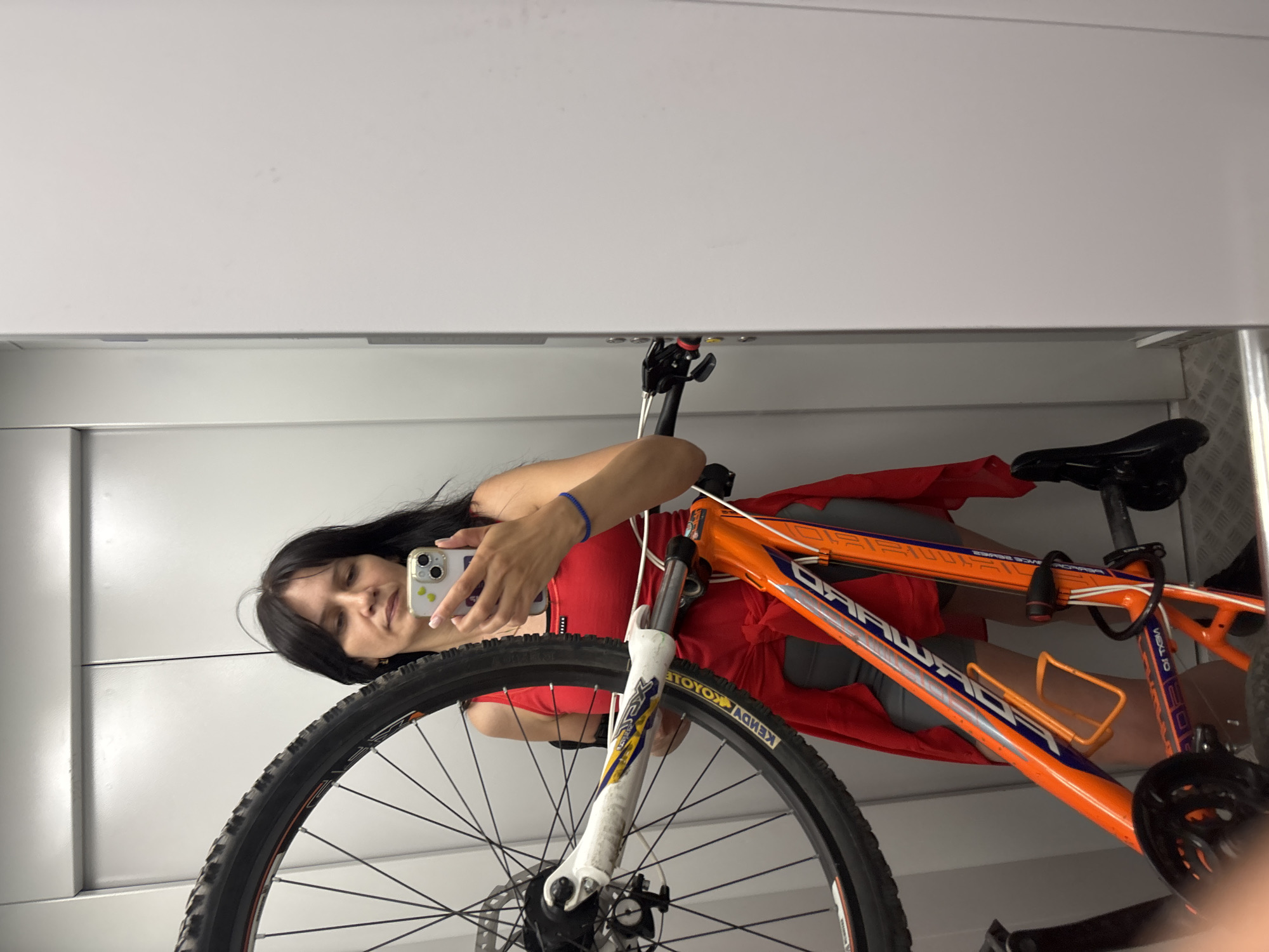 In a dimly lit elevator, a woman with dark hair is holding an orange and white mountain bike. She is wearing a red tank top, grey shorts, and a red garment draped over her shoulders. She is using her mobile phone to take a selfie, and her reflection is visible in the elevator's mirrored wall. The elevator's interior is metallic grey and white, with a checkered floor. The woman is standing in the city of Chaikovsky, Russia.