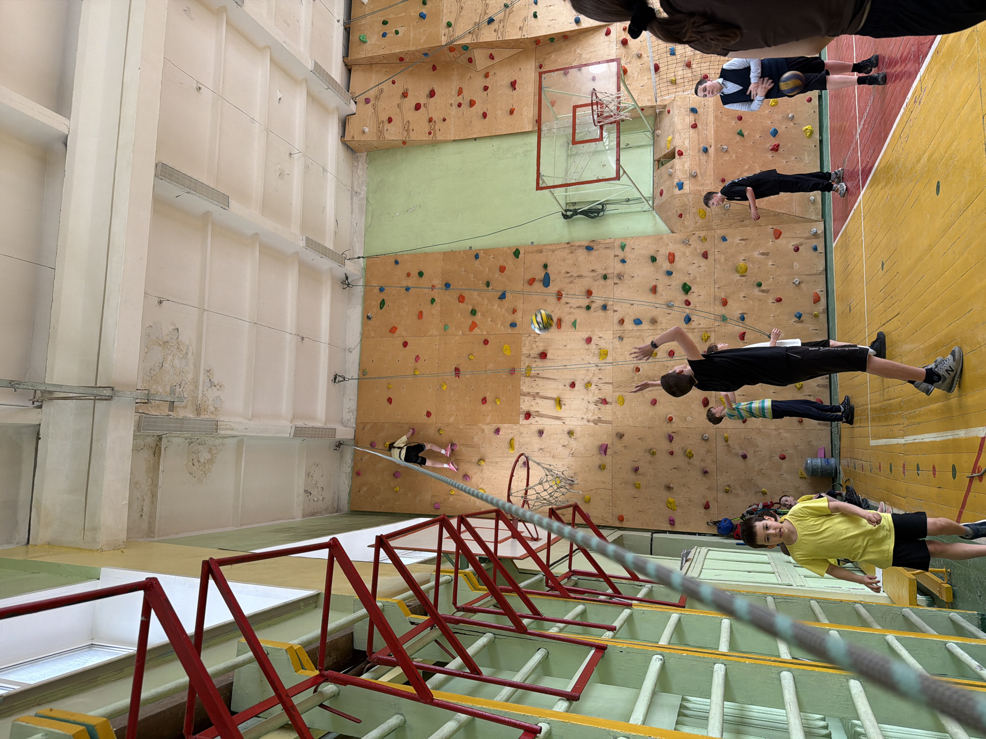 The image captures an indoor sports facility with a prominent climbing wall adorned with colorful handholds and a basketball hoop. Several individuals are present, some positioned near the climbing wall, while others are on the gymnasium floor.
The foreground contains red and yellow climbing equipment. There are eight people scattered throughout the scene. One person appears to be climbing, suspended by a rope. One individual is holding a volleyball.
The facility exhibits high ceilings, and the walls appear to be in varying states of repair, with some sections showing signs of wear.
While the image's location in Izhevsk, Russia, cannot be definitively confirmed, the details suggest an indoor sports facility.