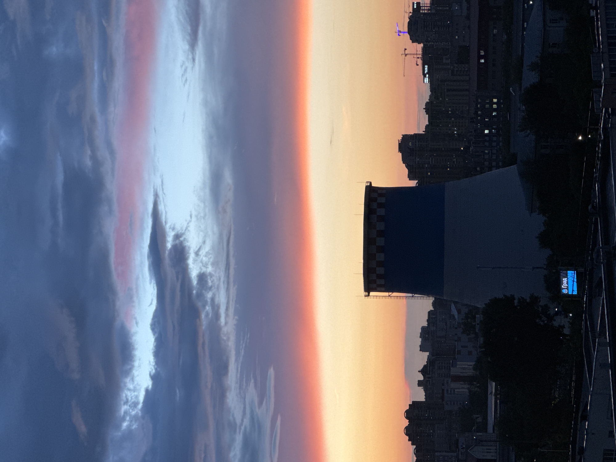 This vertical, medium shot captures a twilight view of a city skyline, likely Moscow, Russia, dominated by a large industrial cooling tower in the foreground. The sky displays a gradient of colors, transitioning from deep blue and grey clouds at the top to a vibrant band of orange and pink hues, indicative of sunset or sunrise. The cooling tower, a prominent structure, is cylindrical with a wide base and tapers towards the top, featuring a checkered pattern of dark blue and white near its rim.
Below the sky and behind the cooling tower, a dense city skyline is visible, characterized by numerous buildings of varying heights and architectural styles, many of which are illuminated with interior lights, suggesting it is evening or dusk. Trees and other foliage are scattered throughout the urban landscape, some silhouetted against the brighter sky. Construction cranes are also visible among the buildings, hinting at ongoing development within the city. In the lower portion of the frame, a blue LED sign with white Cyrillic text reading "Град" is illuminated, adding a modern touch to the scene. The overall atmosphere is one of urban twilight, with the industrial structure of the cooling tower contrasting with the natural beauty of the sky and the bustling city below.
