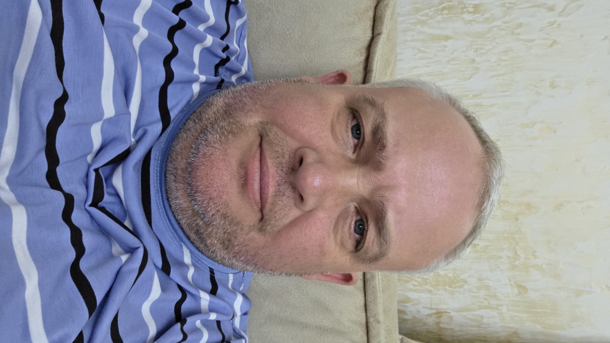 This media file is a close-up, head-and-shoulders portrait of a middle-aged man. He has fair skin, a light beard, and graying hair. His eyes are blue and he is looking directly at the camera with a neutral expression. He is wearing a blue and white striped shirt with black wavy stripes. The man is lying down, as indicated by the tilt of the image, on what appears to be a couch or bed. The background is a textured wall in a light, creamy color. The lighting suggests it is daytime, possibly indoors, as there are no strong shadows. The image is taken from a slightly low angle. There is no visible text. The location context is Batumi, Georgia.