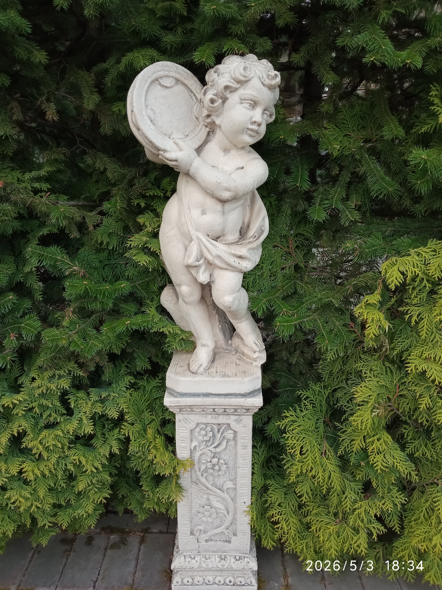 A statue of a child holding a round object stands on a pedestal. The statue and pedestal are light in color. The statue is positioned in front of a green, leafy plant. The date 2026/5/3 18:34 is printed in the lower right corner of the image. The image was likely taken in Krasnoyarsk, Russia based on the surrounding vegetation.