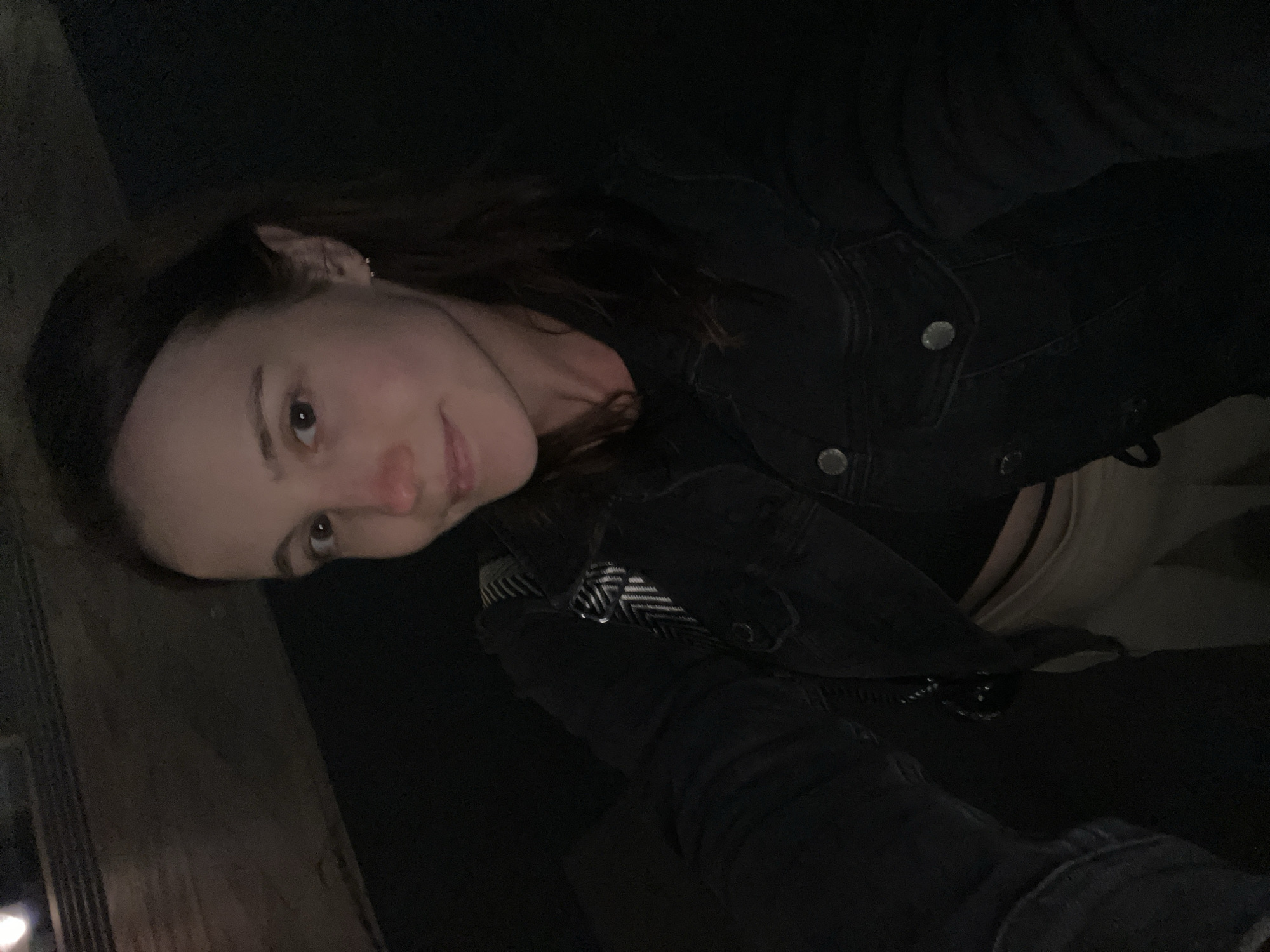 The image is a selfie taken at night, with the subject, a woman with brown hair and fair skin, looking at the camera. She is wearing a dark denim jacket over a black top and light-colored pants. She has a small earring in her left ear. The background is dark and appears to be outdoors, possibly in an urban setting. The lighting is dim, with some light sources visible in the upper left corner of the image, suggesting artificial lighting. There is no specific identifiable landmark or context to pinpoint the location to Novokuybyshevsk, Russia.