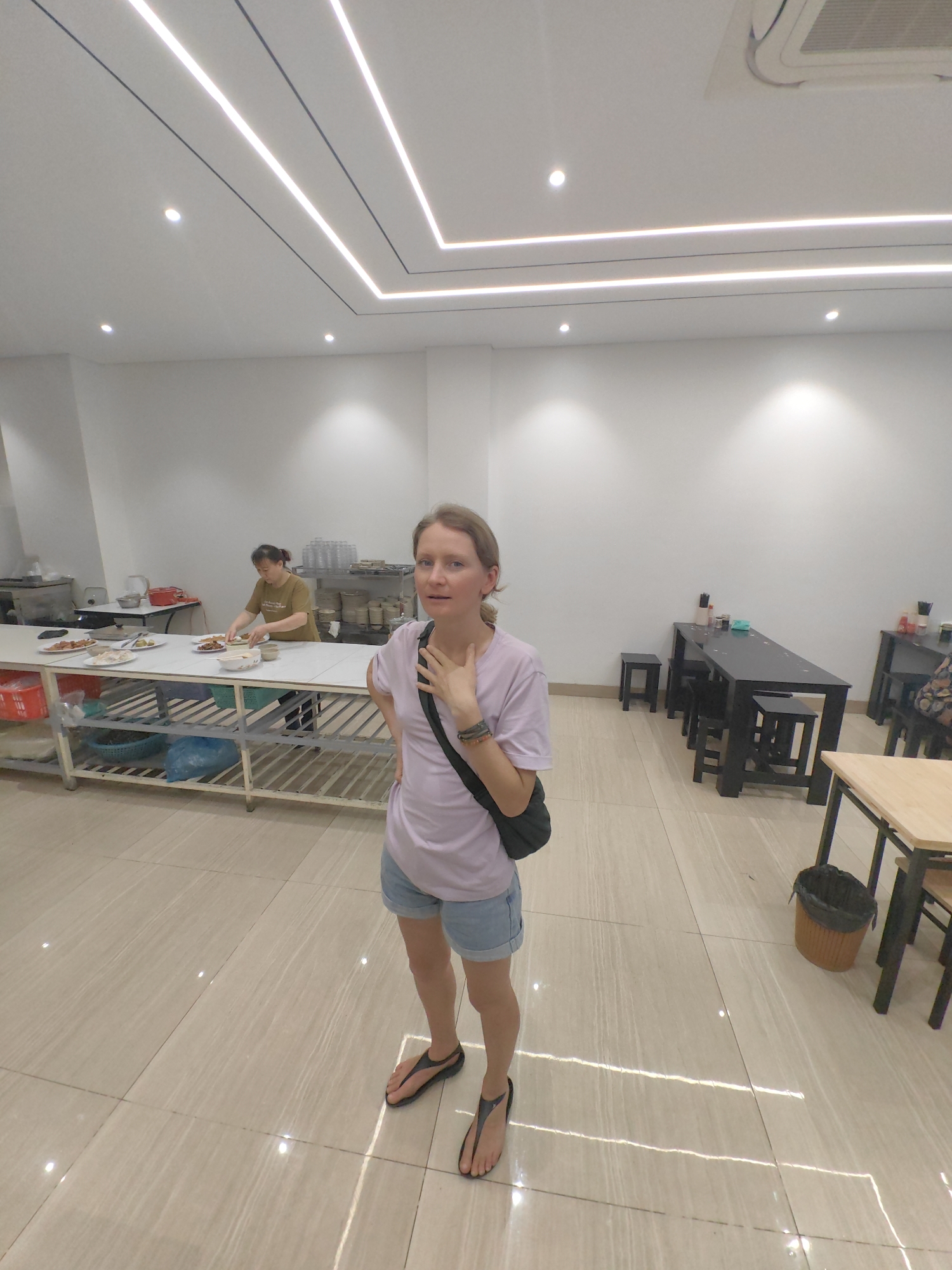 The image shows a woman in a restaurant in Hong Gai Ward, Vietnam. She is standing in the foreground, wearing a light purple t-shirt, blue denim shorts, and black flip-flops. She has a black bag slung over her shoulder.
Behind her, a woman is working at a table laden with food, possibly preparing or serving dishes. There are stacks of plates and other kitchen items visible.
Several black tables with stools are placed near the back of the room. To the right, a light wooden table with chairs is present. A trash can sits near this table. The floor is tiled in a light color.
The room is well-lit with modern, linear lighting fixtures on the ceiling. The walls are painted white, and the overall impression is of a clean and well-maintained restaurant environment.
