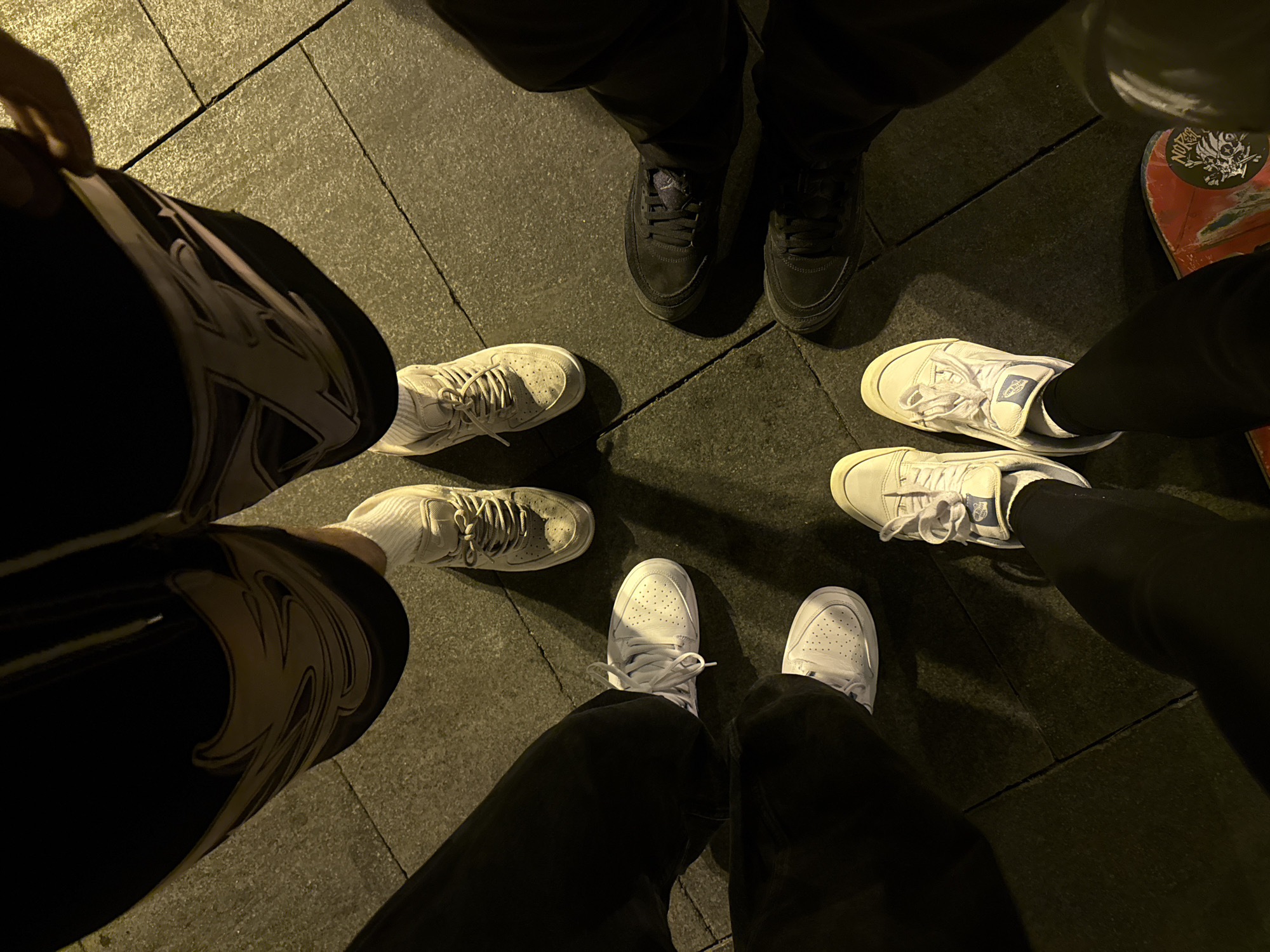 The image shows a group of six people standing in a circle, their feet and lower legs visible. The ground is paved with rectangular tiles, suggesting an urban setting. There are six pairs of shoes visible: one pair of black sneakers, two pairs of white sneakers, one pair of off-white sneakers, one pair of white sneakers with blue details, and one pair of sneakers that cannot be fully seen due to being only partially in the frame with a dark-colored skate deck near the legs of the person. The clothing worn by the individuals is primarily dark-colored, with black pants. No faces are visible. The photo is taken at night, under artificial light, causing deep shadows. No specific details indicate a location in Barcelona, Spain.