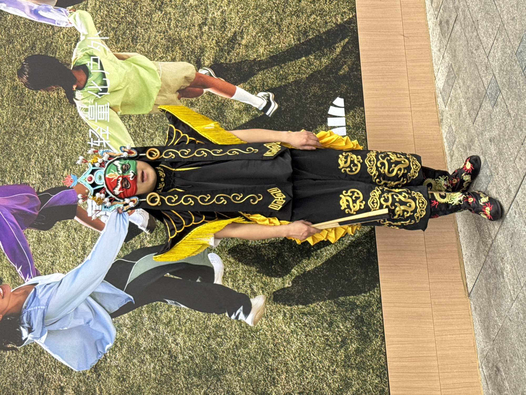 The image depicts a person wearing a Chinese opera costume and makeup, lying on a large advertisement poster on the ground. The costume is primarily black with gold embroidery, and the makeup includes green, red, and white face paint. The poster beneath the person depicts other people in various poses on a grassy field. The ground surrounding the poster is paved with rectangular tiles. The image was taken in the city of Jinjiang District, China.