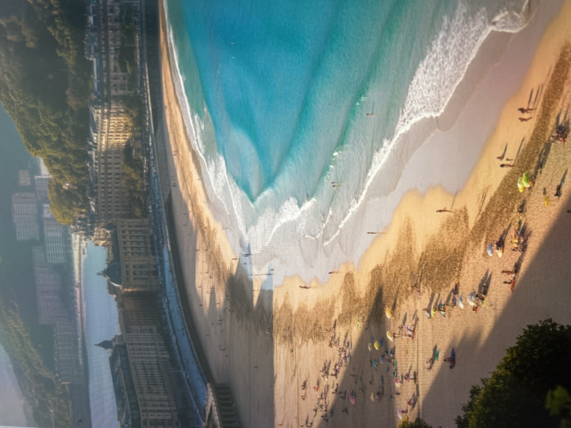 This is an aerial, high-angle view of a bustling crescent-shaped sandy beach and coastline, seen as if rotated approximately 90 degrees counter-clockwise from its natural horizontal orientation.
The scene features a wide, golden sand beach curving around a bay. The water transitions from a light turquoise near the shore to a deeper blue further out, with white foamy waves breaking gently onto the sand. Numerous people are scattered across the beach, some closer to the water's edge, and many further back, often gathered around colorful beach umbrellas. Long, distinct shadows of people and structures are cast across the sand, suggesting a low sun angle, indicative of either early morning or late afternoon. A few small figures can be seen in the clear water, possibly swimming or wading, and what appear to be small boats or kayaks are visible in the bay.
Along the curve of the bay, behind the beach, runs a promenade or walkway, bordered by a line of multi-story buildings. These buildings exhibit classical European architectural styles, with some featuring prominent domes or ornate rooftops. Beyond the urban strip, lush green hills covered in dense vegetation rise up, enclosing the bay. In the distance, further parts of the city with more residential or commercial buildings can be glimpsed.
The overall impression is one of a scenic and lively coastal city with a popular beach where people are enjoying a sunny day. No visible text is present in the image.
