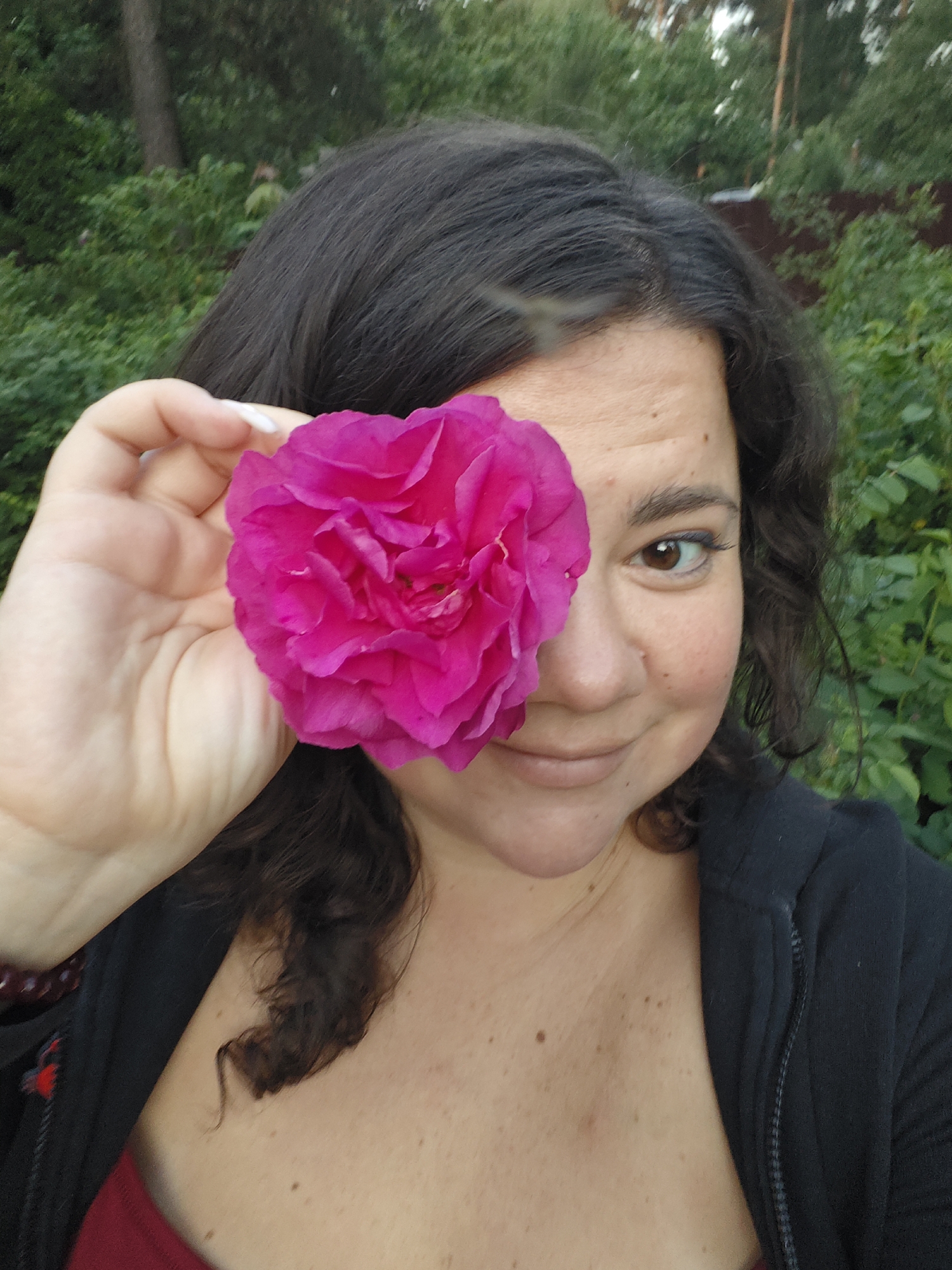 A close-up portrait shows a woman holding a vibrant pink rose over her left eye, partially obscuring her face. She has dark, wavy hair and light brown skin with scattered freckles or moles. Her right eye, a warm brown, is visible and looking directly at the viewer, accompanied by a subtle, pleasant smile.
Her left hand, with white-painted fingernails, holds the multi-petaled rose. She is wearing a dark, possibly hooded, top over a reddish undergarment. A dark red beaded bracelet is visible on her left wrist.
The background consists of lush green foliage, including bushes and trees, suggesting an outdoor, natural setting like a garden or park. The lighting appears soft and natural, indicating it is daytime, possibly late afternoon. No text is visible in the image.