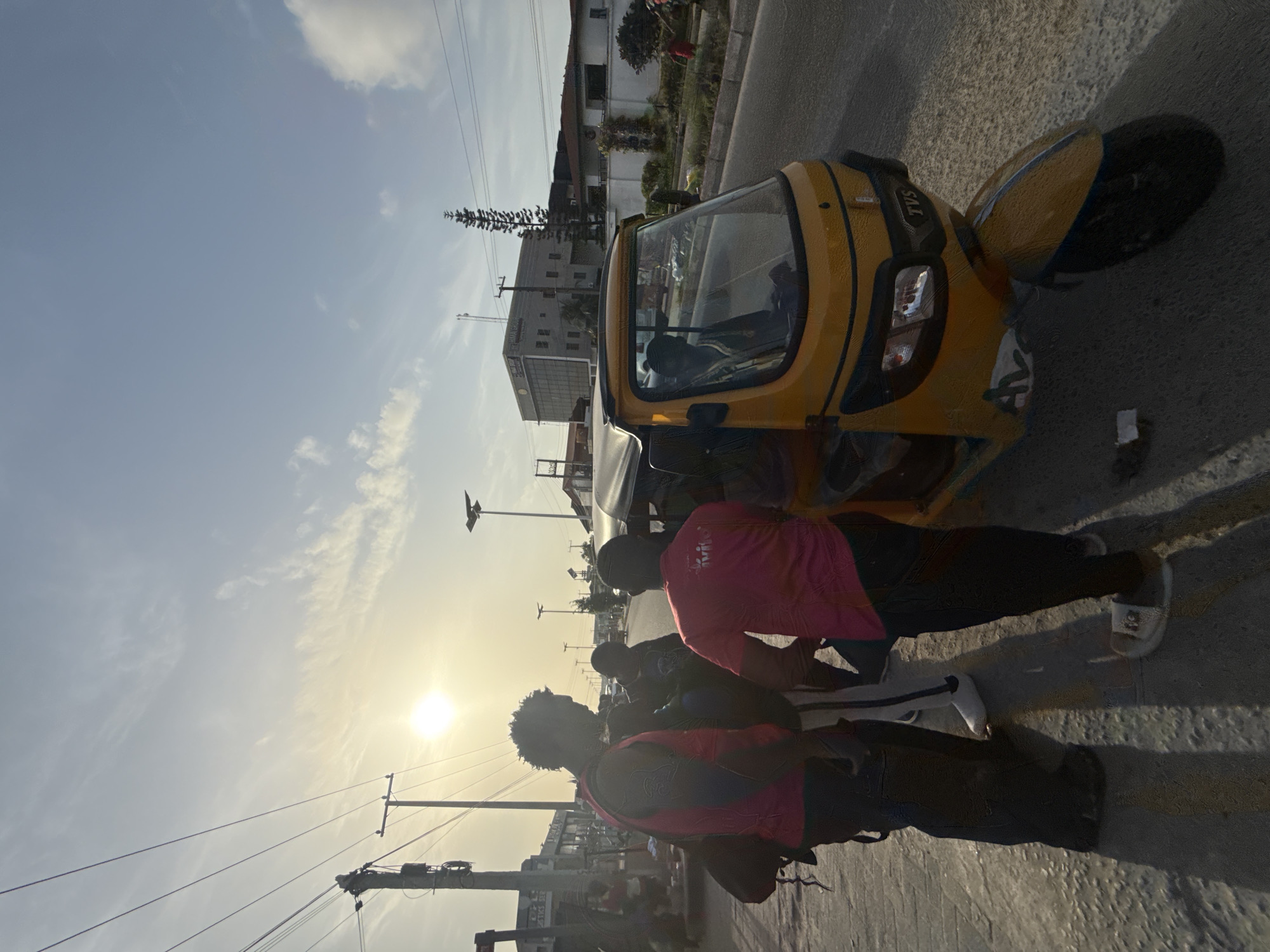 Here's a detailed description of the image:
**Overall Scene:** The image appears to be taken on a street in Ado, Nigeria, during daylight hours, with the sun low in the sky. The angle of the photograph is sideways.
**Subjects & Objects:**
* A yellow "Keke Napep" or auto-rickshaw is prominent in the shot. It looks like a relatively new model with a windshield and side mirrors. The vehicle is parked on the side of the road.
* Two people are standing or interacting near the auto-rickshaw. Both are wearing what appear to be pink shirts or tops.
* Other pedestrians are visible further down the road, but they are not in focus.
* Power lines are visible stretching across the sky, a common feature in many cities.
* Several buildings are in the background.
**Actions & Interactions:**
* The two people are standing by the auto-rickshaw, it looks like they're interacting together.
**Environment:**
* The location is a paved street in what appears to be a populated area.
* There are buildings nearby, indicating an urban or semi-urban setting.
* The presence of power lines further reinforces the impression of an urban environment.
* The sun is shining, creating harsh shadows.