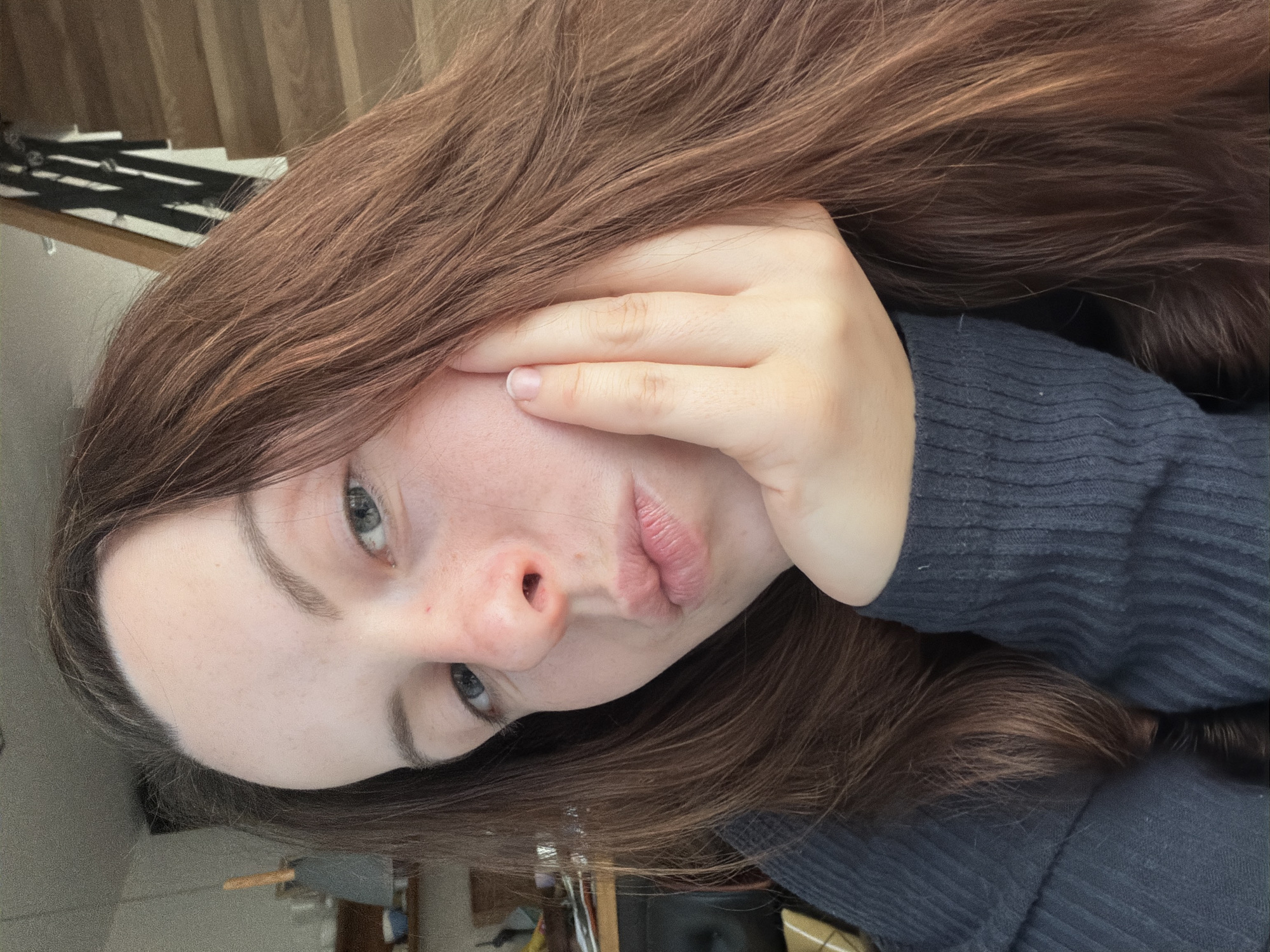 In an indoor setting, a young woman with long, brown hair rests her head on her hand. She is wearing a dark gray sweater. Her hand is propped against her cheek, and her lips are pursed. The background is blurred but suggests a home environment with a wooden staircase and shelves with various items. There is no indication of the location being in Gangloff, Germany.