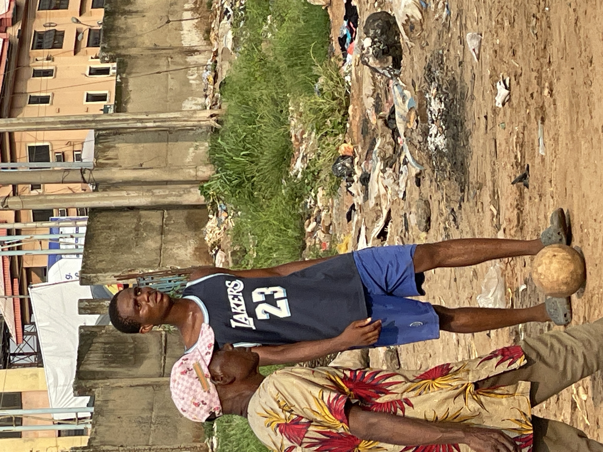 The image depicts an outdoor scene in an unspecified urban or peri-urban area within the city of Unknown, Unknown. The ground is composed of dry, light brown dirt or sand, heavily littered with various discarded materials including plastic bags, fabric scraps, and general debris. Patches of green grass and weeds grow amidst the refuse. In the mid-ground, a small circular area of ash and charcoal indicates a past fire pit.
Two men are present. On the left, partially cropped, an older man with dark skin walks rightward, wearing a pink and white checkered baseball cap with a brown rectangular patch, a beige short-sleeved shirt patterned with red and yellow tropical leaves, and light brown trousers. His right arm and hand are visible.
To his right, a younger man with dark skin stands still, facing rightward with a pensive expression, looking slightly upwards. He wears a dark blue basketball tank top with "LAKERS" and "23" printed in white on the front, blue athletic shorts, and grey slip-on shoes. A light brown, spherical object resembling a makeshift ball rests on the ground near his feet.
In the mid-background, a weathered, grey concrete wall runs horizontally, separating the immediate foreground from further structures. Beyond this wall, various multi-story buildings with light brown or beige facades and dark windows are visible under a clear sky. A large white tarp or banner, with a partial black letter "g." visible, covers a structure to the left of the background buildings. Several vertical utility poles are also visible.