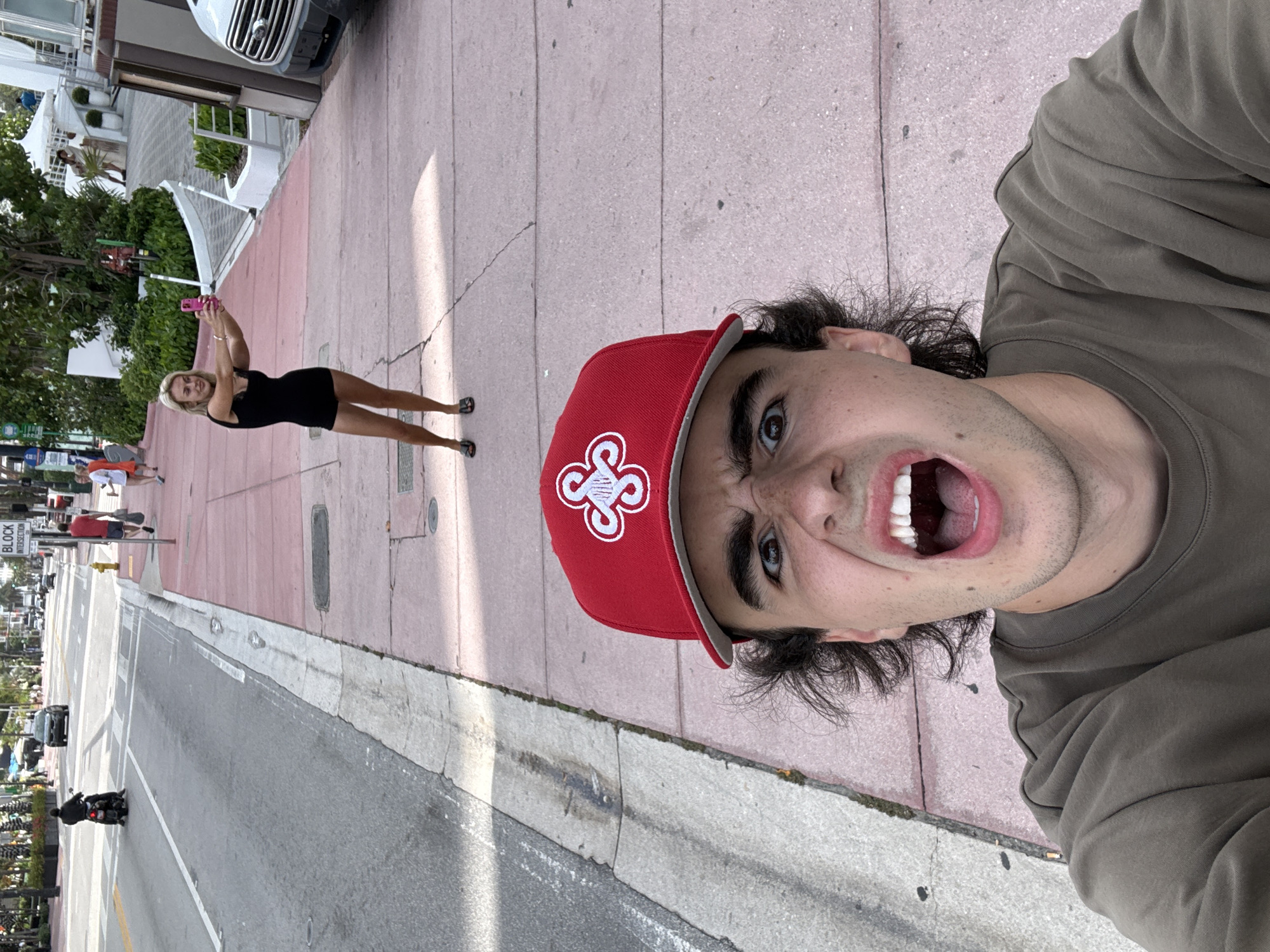 A street-level, eye-level photograph shows a light-skinned male with brown hair and a red baseball cap with a white logo. He is wearing a gray t-shirt. His mouth is wide open, and he is looking upward. Behind him, a light-skinned woman with blonde hair is standing on the sidewalk, facing toward the buildings. She is holding a pink phone and appears to be taking a photograph. She is wearing a black dress and black sandals. The sidewalk is pink and gray. A street is visible with a motorcycle riding on it. A block intersection sign is visible. The location is Miami Beach, United States of America.