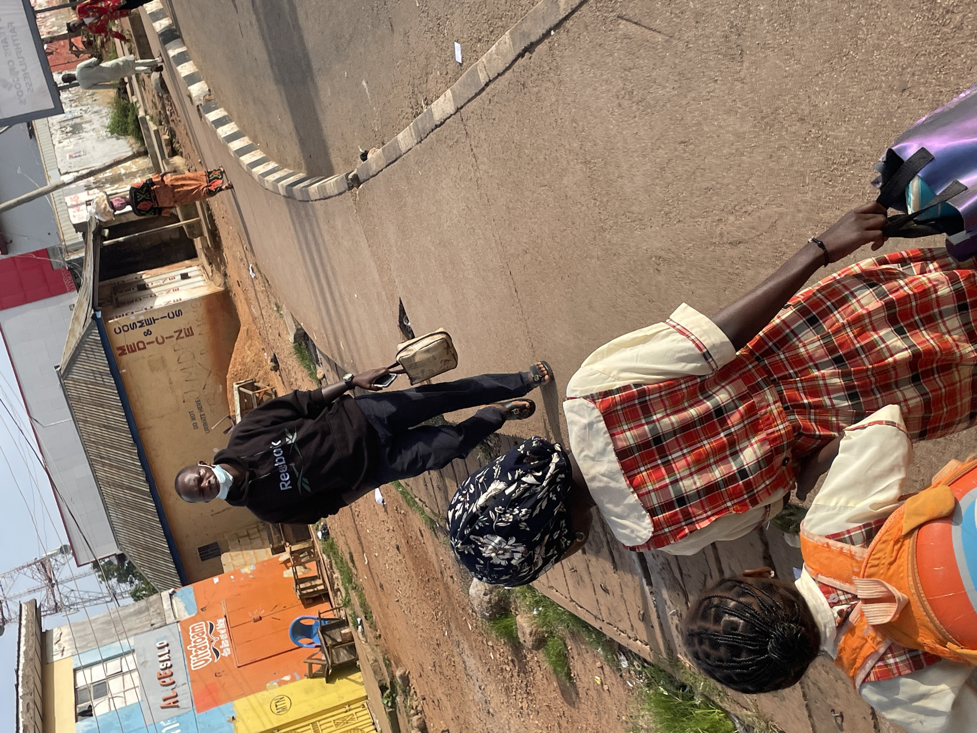 In Jos, Nigeria, a man wearing a black Reebok hoodie and a face mask walks on a sidewalk next to a street. He carries a light brown tote bag in his right hand. In the foreground, a young girl wearing a red and white plaid dress and a white shirt, with an orange backpack, walks away from the camera. Her hair is styled in braids. Another person wearing a dark headscarf with a floral pattern is also walking on the sidewalk. In the background, a woman in an orange dress carries a bundle on her head and walks on the street. Buildings with colorful facades and corrugated roofs line the street. A large communication tower is visible in the distance. The ground is a mix of dirt and concrete.