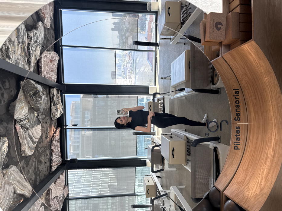 A woman in black athletic wear takes a selfie in a pilates studio. Large windows reveal a city skyline. Pilates equipment is arranged throughout the bright space. The studio features a textured wall with embedded stones. A curved wooden structure with the words "Pilates Sensorial" is visible, adding a design element to the studio.