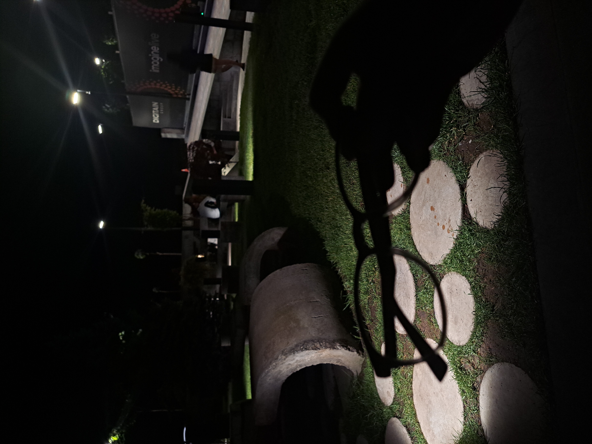 The image depicts an outdoor scene in Yerevan, Armenia, at night. A partial view of a person holding eyeglasses is in the foreground, casting a large shadow. In the midground, there is a grassy area with round stepping stones. Also in the midground, there are concrete cylindrical structures, seemingly decorative, and partially illuminated. In the background, there is a banner with the word "imagine live" and "DIGITAIN". People can be seen in the background. There are streetlights visible as well. The overall lighting is dim, with artificial light sources causing reflections.