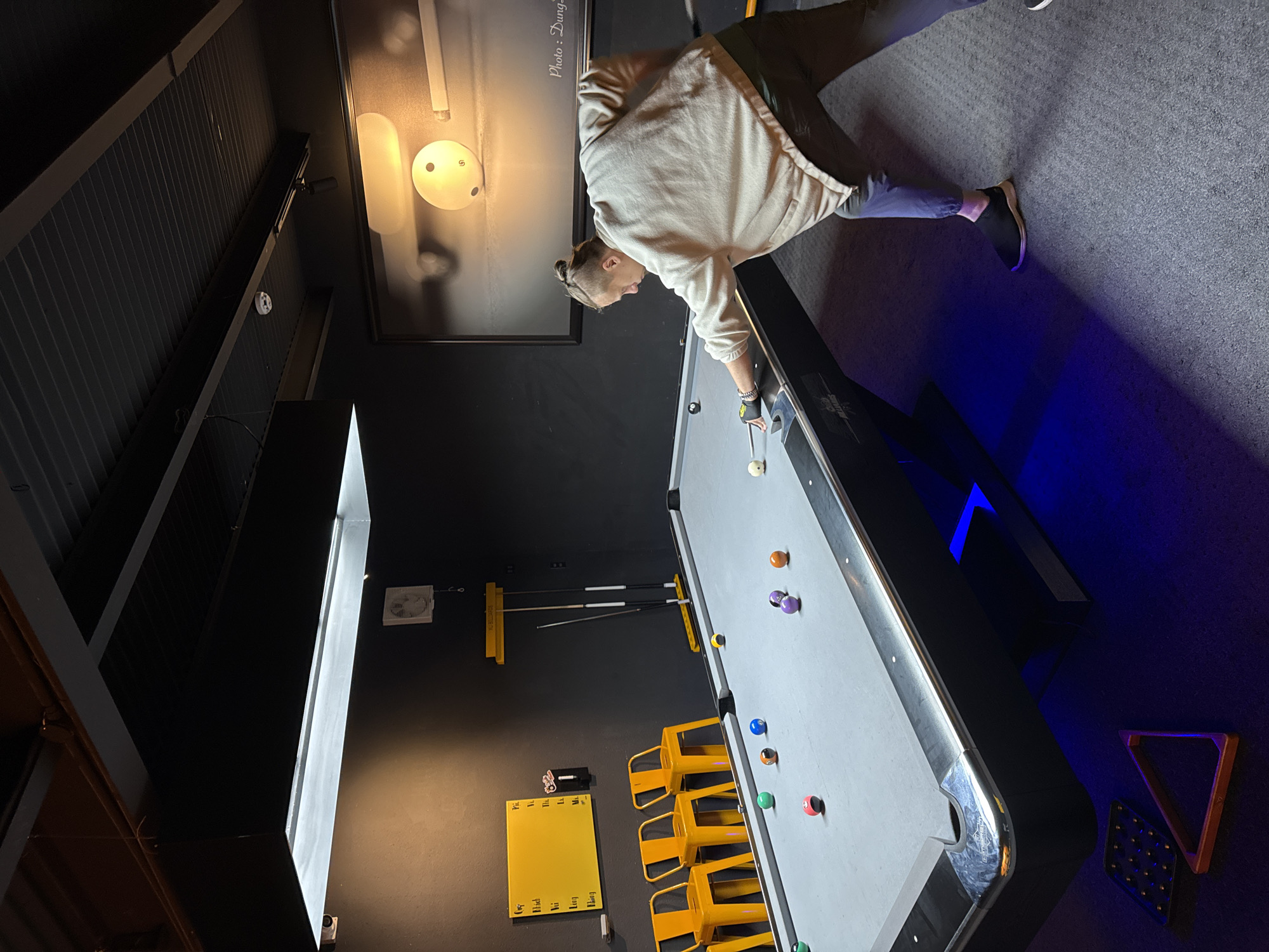 A person is playing billiards in an indoor recreational area. The setting includes a billiards table with balls positioned on it, surrounded by yellow stools. A yellow scorecard and pool cues are mounted on the wall. A framed black and white photograph hangs on the wall, partially obscured by glare. The environment is lit with ambient blue light. The location is not specified, but based on clues, it's possibly within Phường Xuân Hương - Đà Lạt, Vietnam.