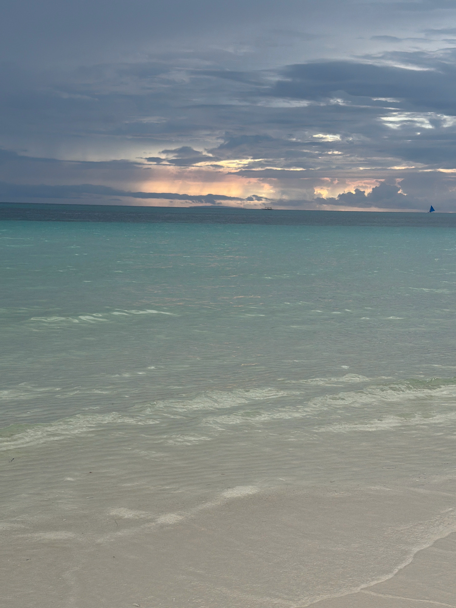 This is a landscape photograph of a beach and the ocean at Boracay Island, Malay, Philippines. The image is dominated by a calm, turquoise ocean that transitions into a sandy beach in the foreground. The sky above is filled with dramatic, cloudy formations, with hints of sunrise or sunset colors peeking through in the distance. A small sailboat with a blue sail is visible on the horizon, along with a distant vessel. The water's surface shows gentle ripples and some lighter areas that suggest sandbars or shallow water. The beach itself is a pale, light brown, with the gentle curve of a wave washing ashore. The overall mood is serene and tranquil.