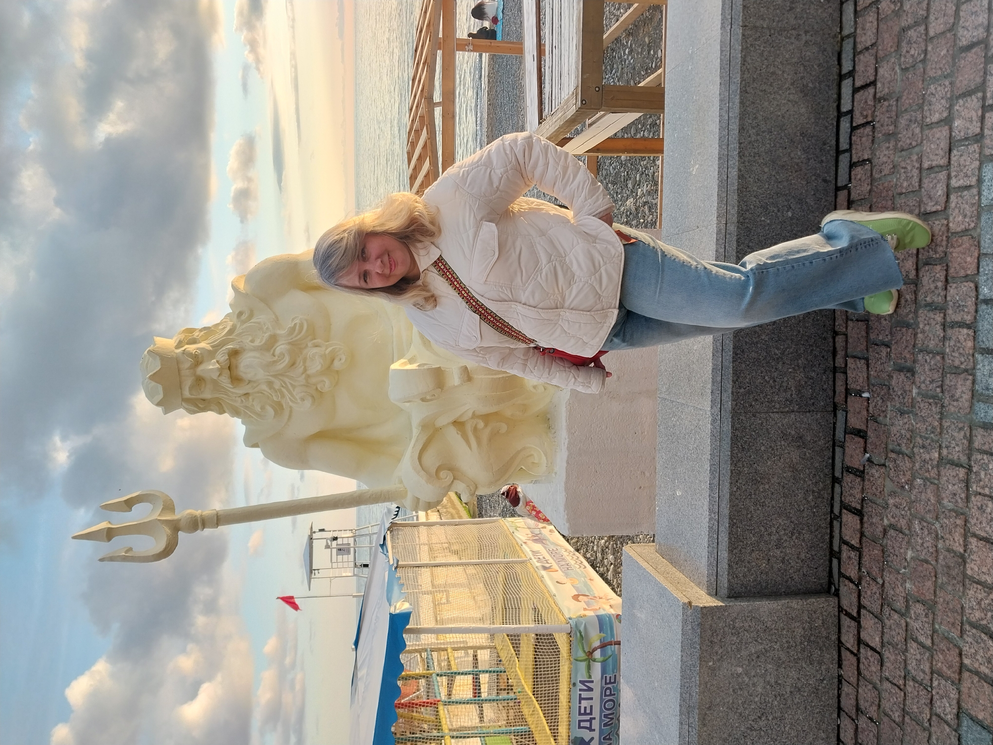 A woman stands next to a sculpture of Poseidon with his trident in Sochi, Russia. She wears a white quilted jacket, jeans, and green shoes. The background includes the sea, a beach, and a small playground. The path is paved with red stones.