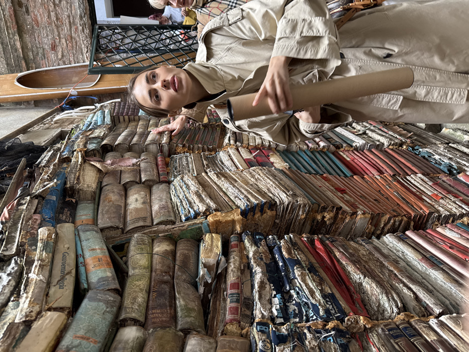 The image depicts a narrow, book-lined passageway within Libreria Acqua Alta in Venice, Italy. The walls on both sides of the passage are constructed entirely from irregularly stacked, aged, and often water-damaged books, forming dense, multi-colored masses from floor to ceiling. In the foreground, a young adult female with light brown hair and a light-colored trench coat stands, holding a light brown cardboard tube in her left hand. Her gaze is directed upwards and to the right, beyond the frame. A second, partially visible female figure, older with light hair, is positioned behind the primary subject to the right. A hand, appearing to be the primary subject's, rests on the book wall to her left. Visible book titles on spines include "Gastronomia" and "Grande Dizionario Enciclopedico." In the upper right background, a segment of a wooden gondola hangs vertically on a brick wall, and indistinct figures are visible through an opening further back. The environment is cluttered and dimly lit, emphasizing the vast collection of books.