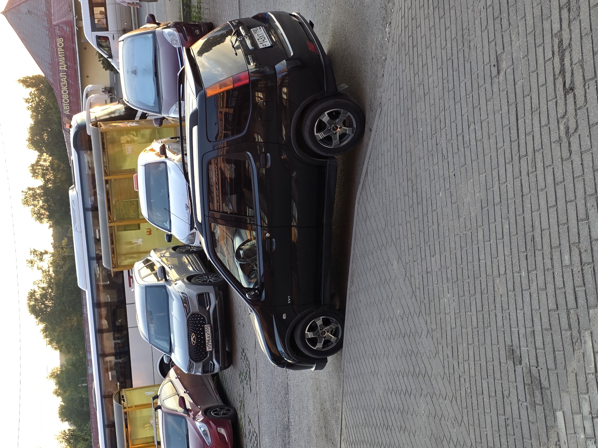An eye-level, medium shot captures a black Suzuki compact SUV parked on a gray brick pavement. The SUV is angled slightly towards the right, with its front passenger door and side visible. To the left of the SUV, a light gray Chery Tiggo SUV is parked, its front end facing forward. Further left, a red Hyundai Accent is parked, its rear quarter panel and side visible. In the background, a long white bus is parked near a bus stop shelter with yellow translucent panels. Above and to the right of the bus, the red roof of a building is visible, with yellow text on it that reads "АВТОВОКЗАЛ ДМИТРОВ". The overall lighting suggests it is daytime, possibly late afternoon, with sunlight casting a warm glow on the scene. The environment appears to be an outdoor urban setting, likely a bus station or parking lot.