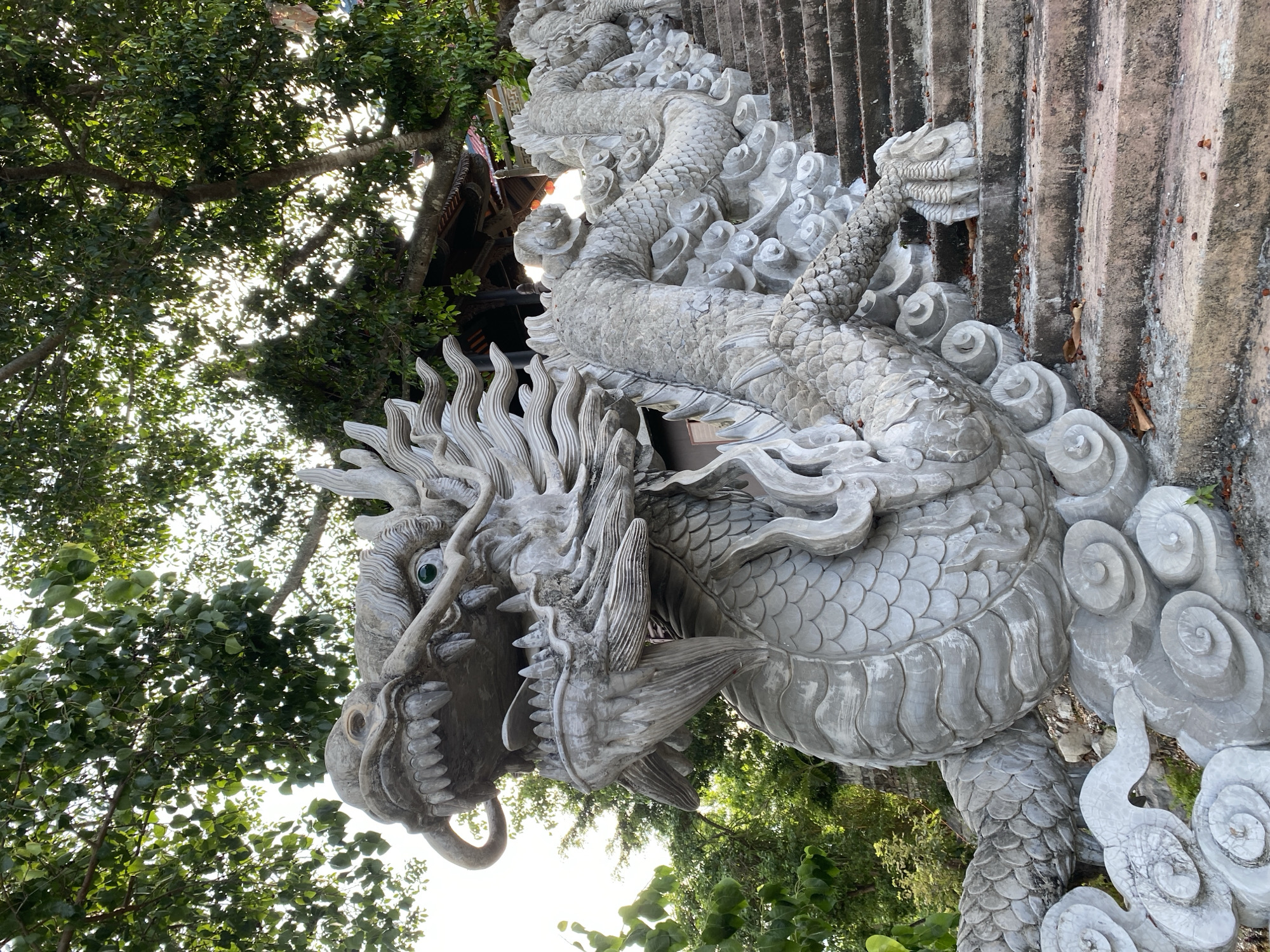 The image shows a stone dragon sculpture adorning the steps of a staircase. The dragon is intricately carved with scales, sharp teeth, and glowing green eyes. It appears to be winding its way up the stairs, which are made of stone and show signs of wear. Lush green trees with sunlight filtering through their leaves form a canopy above and behind the sculpture, suggesting an outdoor setting. The dragon sculpture and the stone stairs suggest this location may be part of a temple, shrine, or a significant landmark. The presence of the dragon, a symbol often associated with power and good fortune in Asian cultures, implies a cultural or religious context. Without further information or visual cues within the image, specifying a precise building or structure beyond "temple or shrine" is not possible.