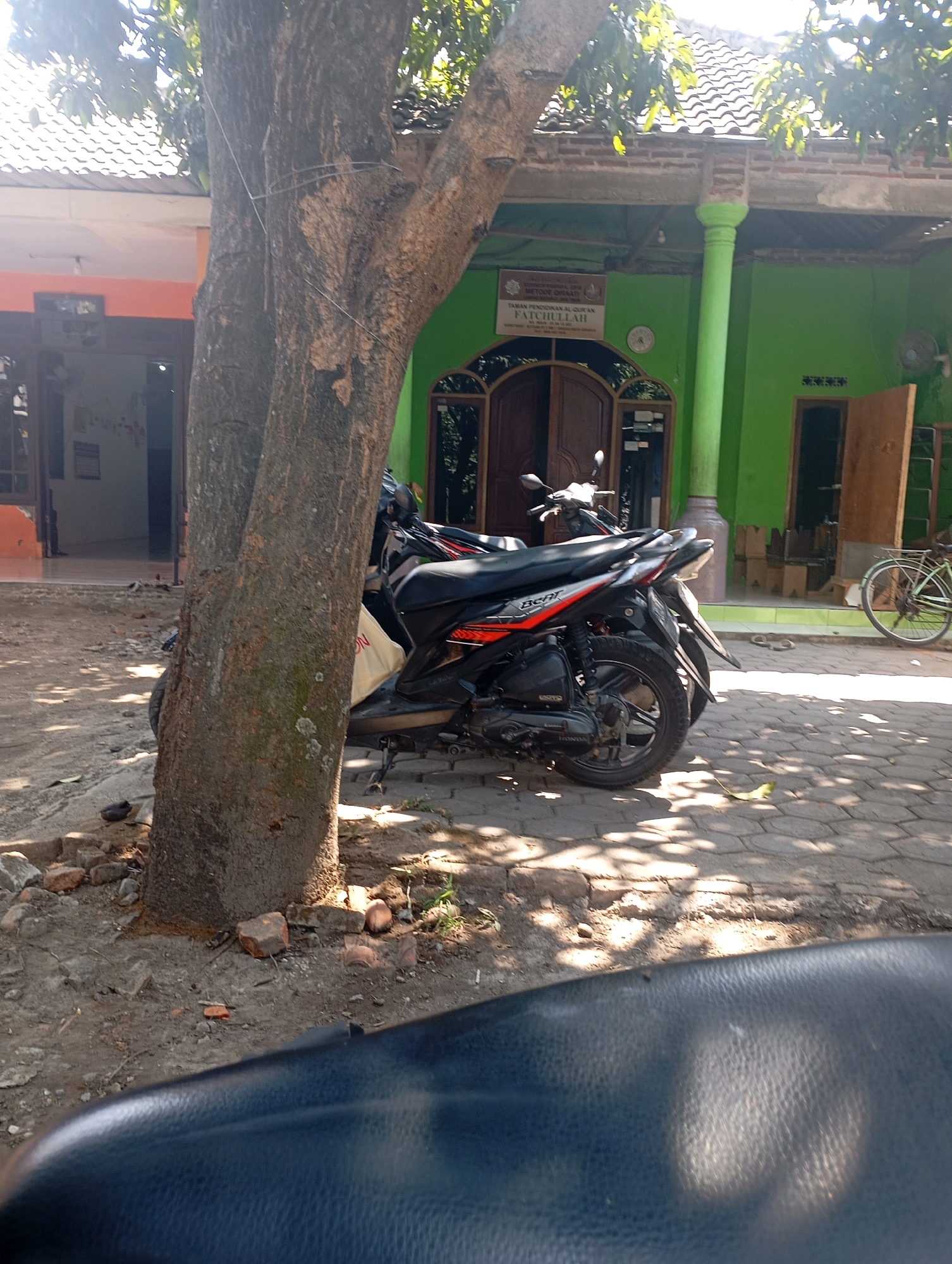 The image depicts an outdoor scene in Ketegan, Indonesia, characterized by a large tree in the foreground, casting shadows on a paved area. To the right of the tree, two motorcycles are parked. One is a black and red scooter with a white bag on its seat. Behind it, another motorcycle is partially visible. Further to the right, a bicycle with a light-colored frame and black wheels is parked against a vibrant green building. The building has a tiled roof and features multiple doors and windows. A sign with text is mounted on the green wall. The ground surrounding the tree is a mix of dirt and scattered bricks. In the foreground, the dark, textured surface of what appears to be a motorcycle seat is visible, suggesting the image was taken from a seated perspective. The lighting indicates it is daytime, with sunlight filtering through the tree's foliage.