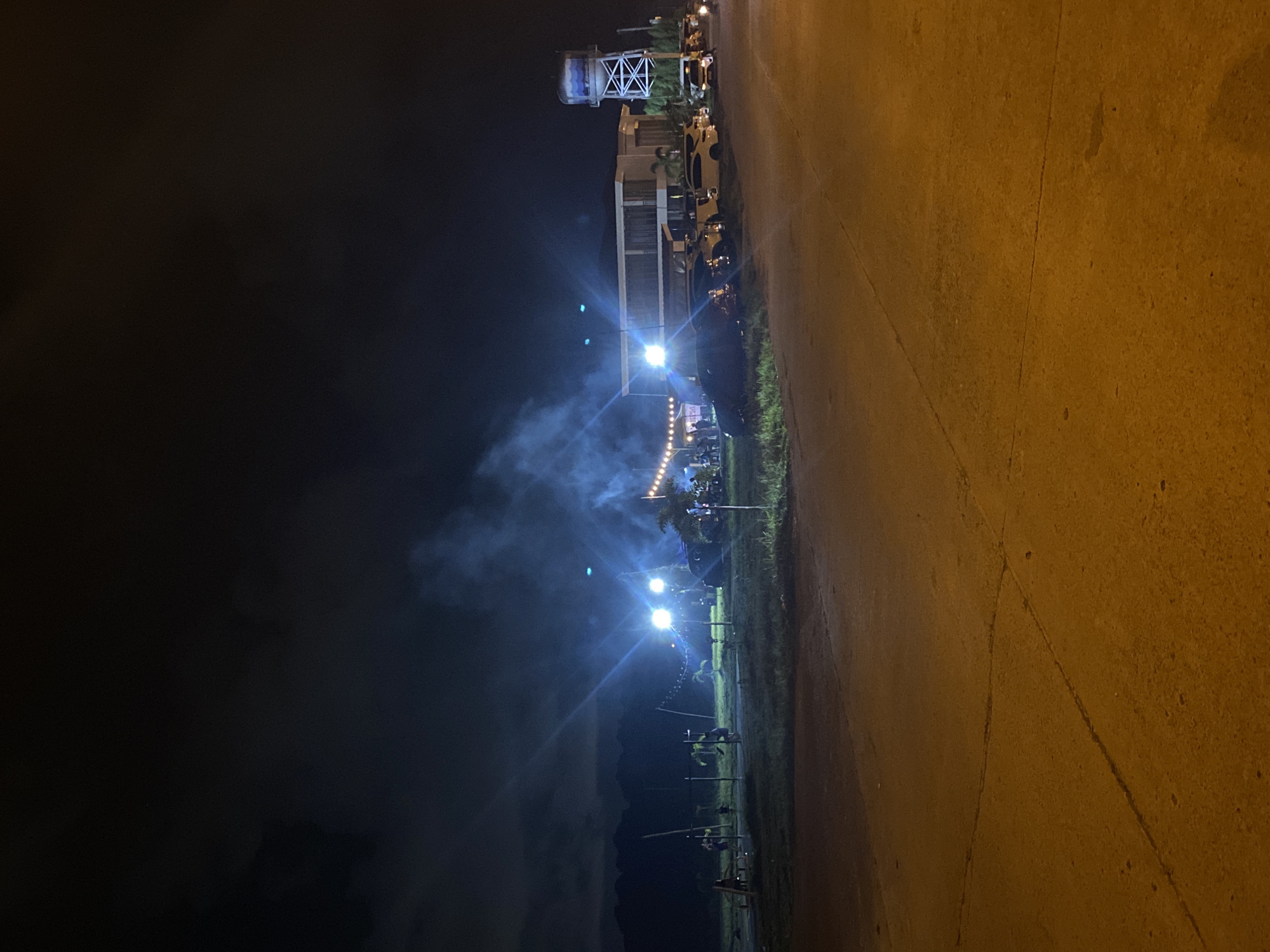 The image depicts an outdoor gathering at night, likely a social event, in Davao City, Philippines. In the foreground, a paved area, possibly a road or parking lot, is illuminated by warm-toned streetlights, showing cracks and debris. Beyond this, a grassy field is dotted with a few palm trees and some rudimentary exercise equipment. Several people are visible scattered across this area, some appearing to be socializing or engaged in activities.
Further back, a modern, multi-story building with a flat roof serves as a backdrop. In front of this building, a row of vehicles, including cars and SUVs, are parked. Strings of decorative lights are strung across the area, illuminating the gathering. A prominent water tower with a blue water tank on a white lattice structure stands to the right of the building, a common sight in many Philippine cities and municipalities. Wisps of smoke or fog are visible, illuminated by bright spotlights, adding to the ambiance. The overall scene suggests a lively event taking place in a semi-developed area, possibly on the outskirts of the city, with mountainous terrain faintly visible in the dark background.