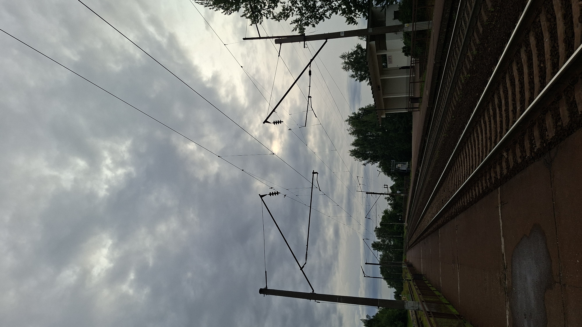 This image depicts a railway track at a train station, likely in Забалоцкі сельскі Савет, Belarus. The perspective is low-angle, looking up towards the sky and along the tracks. The tracks themselves are the primary focus in the foreground, running diagonally from the bottom right towards the upper left of the frame. They are made of ballast and steel rails, showing the standard construction of a railway.
Above the tracks, a complex network of overhead electrical wires and supporting poles stretches across the frame. These are typical of electrified railway lines. The sky is overcast with dramatic, heavy clouds, suggesting a cloudy or potentially stormy day. The sun is partially visible, creating a soft glow through a break in the clouds.
In the background, a small, modern-looking building with a flat roof and a sign is visible to the left of the tracks, suggesting it might be a station building or waiting area. Trees and greenery are present on either side of the tracks and behind the building, indicating a semi-rural or suburban setting. The time of day appears to be late afternoon or early evening, given the quality of light and the cloud cover. No people are visible in the frame, and there is no discernible activity. The overall mood is somewhat dramatic due to the sky, but also mundane, representing a typical railway scene. A small, illegible sign is visible on one of the poles.