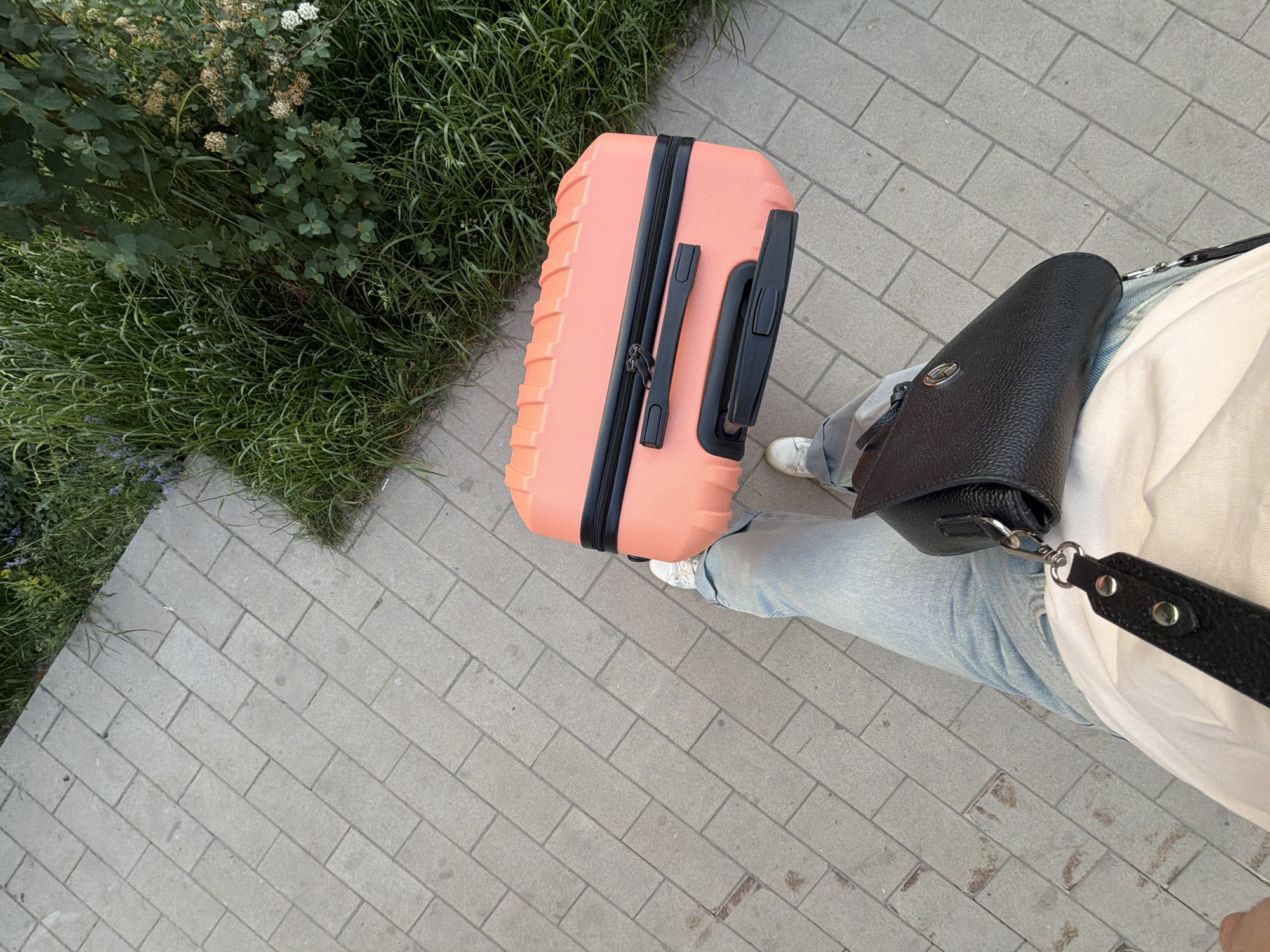 An outdoor, eye-level, medium shot captures a person from the knees down, standing on a grey paved walkway. The person is wearing light blue jeans and white sneakers. A small black shoulder bag with a silver clasp is slung across their body. In their left hand, they are holding a small, bright orange suitcase with a black zipper and handle. To the right of the walkway, there is a patch of green grass with various plants and some small white flowers. The lighting suggests it is daytime, possibly late afternoon, with a soft, diffused light. The image appears to be taken in a city park or residential area.