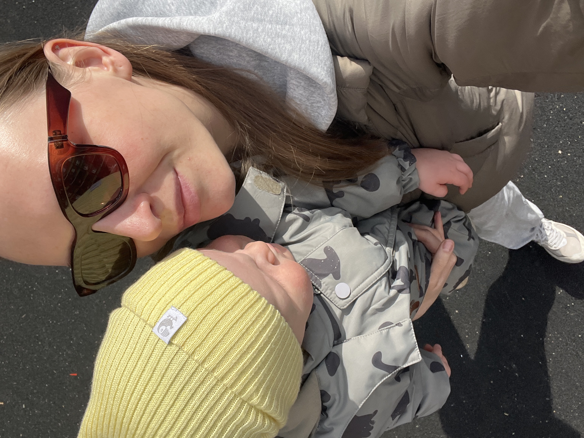 The image depicts a woman holding a baby outdoors. The woman is wearing brown sunglasses and a gray hoodie. The baby is wearing a yellow beanie and a camouflage-patterned jacket. The location is not discernible as Khimki, Russia from the image alone. The ground appears to be asphalt. A small portion of a white sneaker is visible in the bottom right. The lighting suggests it is daytime.