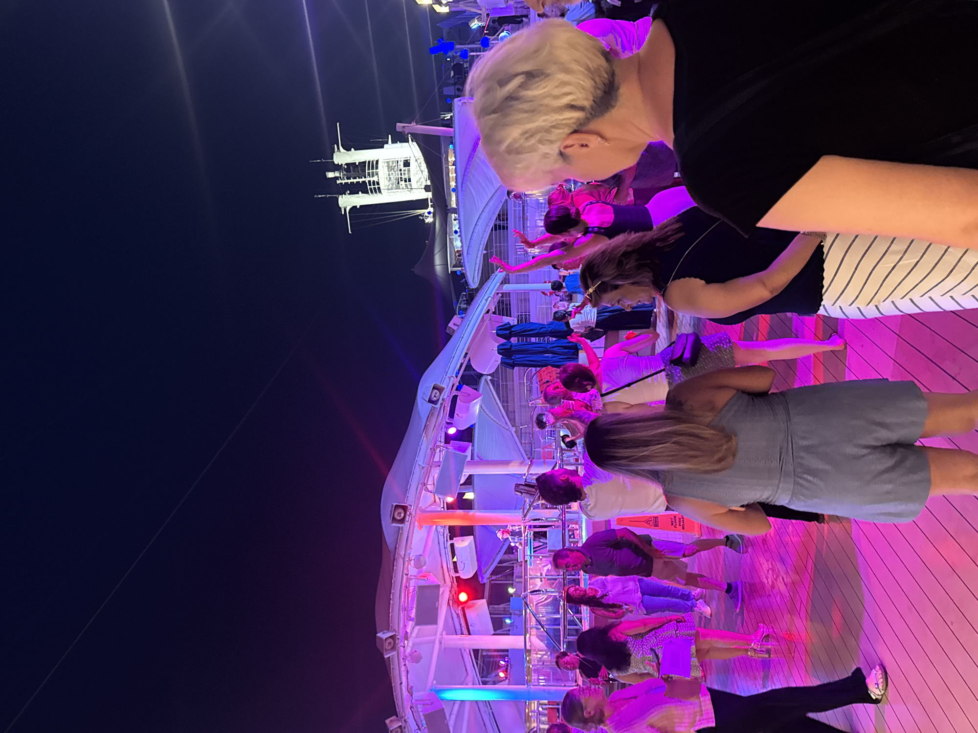 In an outdoor nighttime setting, a group of people are dancing on a wooden deck illuminated by vibrant pink and blue lights. A DJ is visible on a raised platform behind them, and a large cruise ship's superstructure is silhouetted against the dark sky in the background. The image does not depict Salobreña, Spain, or any recognizable urban elements.