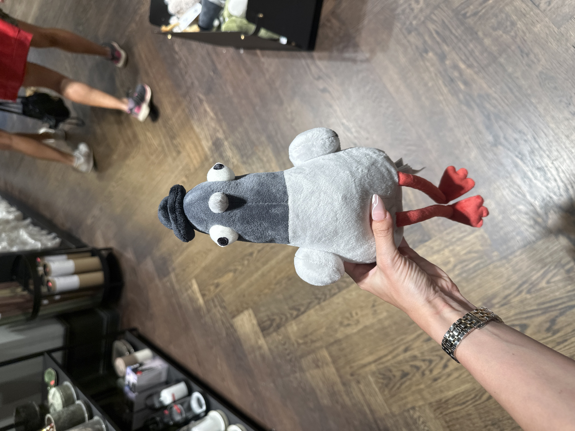 A plush pigeon toy with a black hat is held by a person's hand with a silver watch. The pigeon has large googly eyes, a gray head and body, and red feet. It is being held in front of a wooden floor. In the background, shelves with various items and people walking are visible. The image appears to have been taken inside a store or establishment.