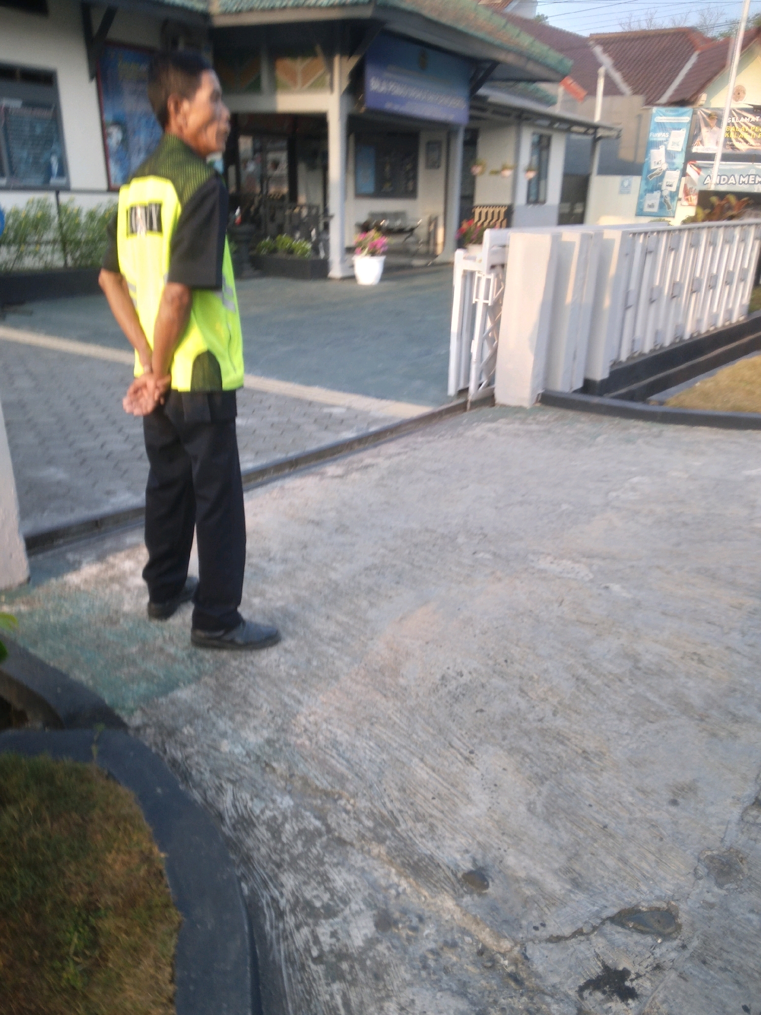 A security guard stands in front of a building in Desa Pamijen, Kecamatan Sokaraja, Indonesia. The guard is wearing a yellow safety vest over a dark uniform and black pants. His hands are clasped behind his back. The building appears to be a public institution with a light-colored facade and a dark-tiled roof. A white fence and a paved walkway are visible in the foreground, with a grassy area to the left. The lighting suggests it is either early morning or late afternoon.