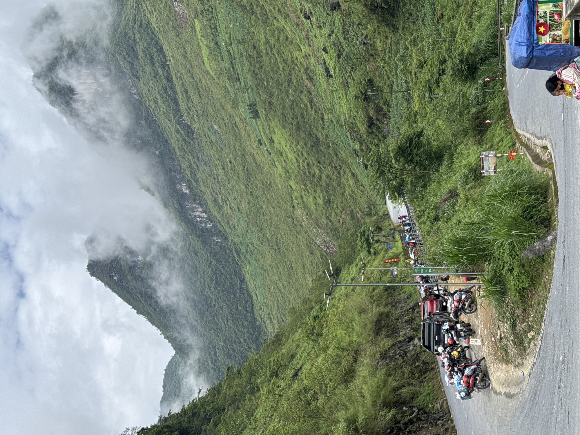 The image shows a steep, green, mountainous landscape, partly shrouded in clouds, in Yên Minh Commune, Vietnam. A narrow road winds along the edge of the mountain. Several parked motorbikes and a few standing people are visible on a roadside platform. A vendor's stand, covered with a blue tarp, is situated beside the road. The road surface is asphalt. Power lines and poles run along the hillside.
