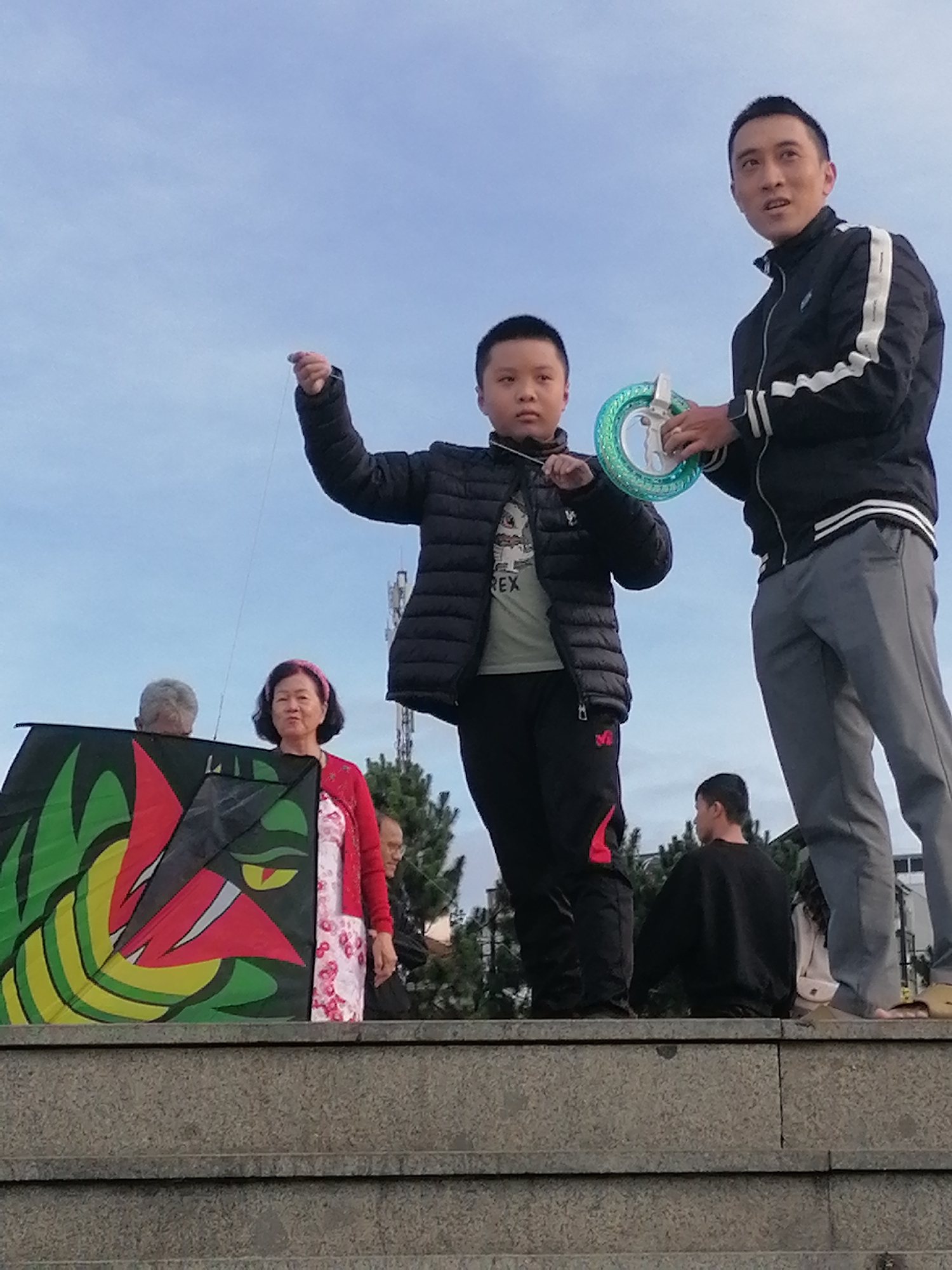 In Da Lat, Vietnam, on a cloudy day, a group of people are gathered on a raised concrete structure. In the foreground, a large, colorful kite designed to resemble a dragon's head is held upright. To the right of the kite, a woman in a red cardigan and a floral dress stands, looking towards the camera with a neutral expression. Behind her, a young boy in a black puffer jacket is focused on flying a kite, holding a string taut. To the right of the boy, a man in a black jacket with white stripes and grey pants holds a kite reel and appears to be talking. Further back, another man in a black sweatshirt is visible. The background features trees and what appear to be telecommunication towers. The overall scene suggests an outdoor activity or gathering in a public space.