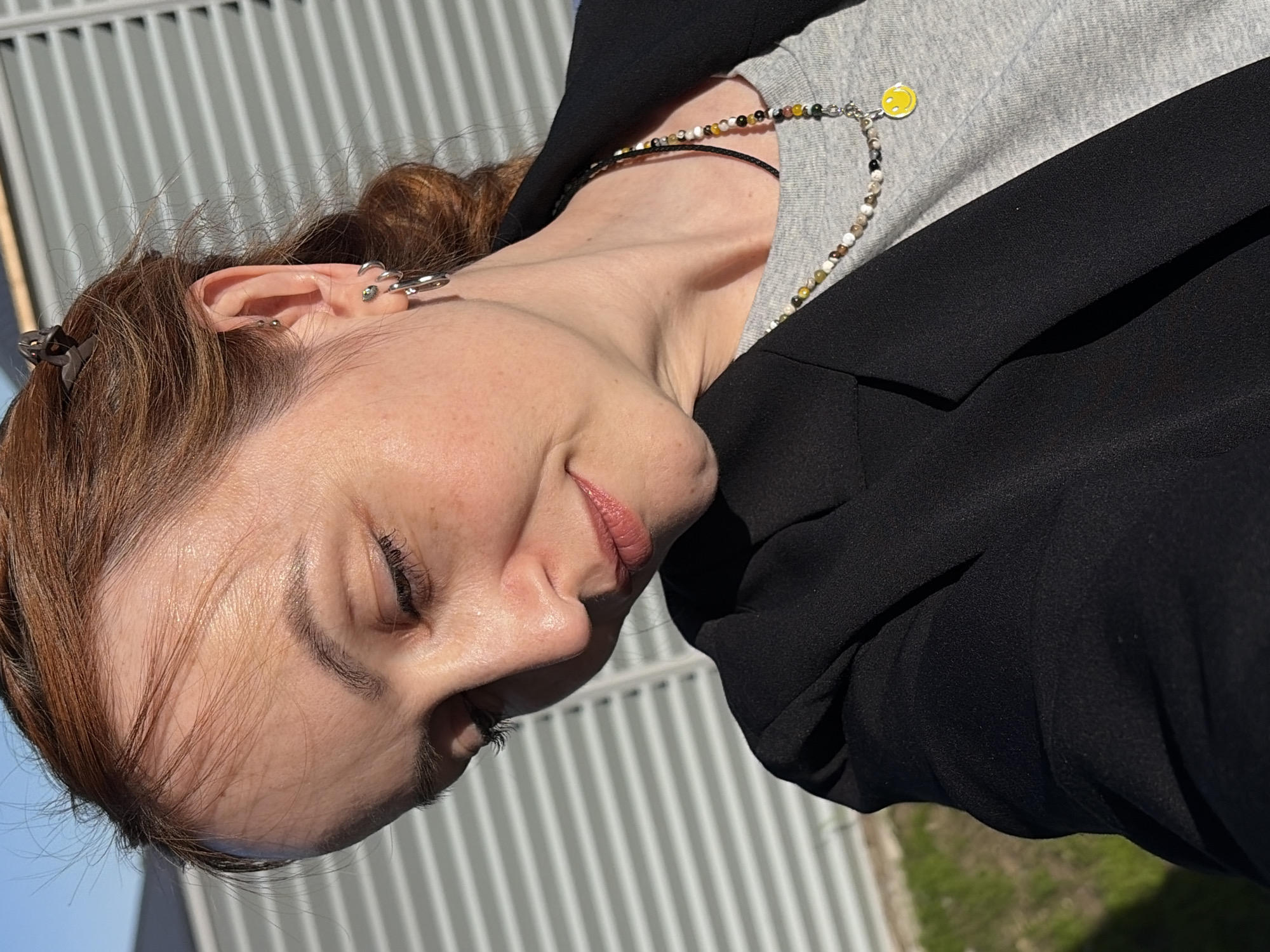 A close-up, angled headshot depicts a fair-skinned individual with light brown hair styled in a bun, a black blazer, and a grey shirt. The individual has multiple piercings on the upper ear. Two necklaces are visible, one with a small yellow smiley face pendant. The background shows a metallic grate and a glimpse of blue sky and greenery. It is impossible to confirm the location is Chaikovsky, Russia without additional information.