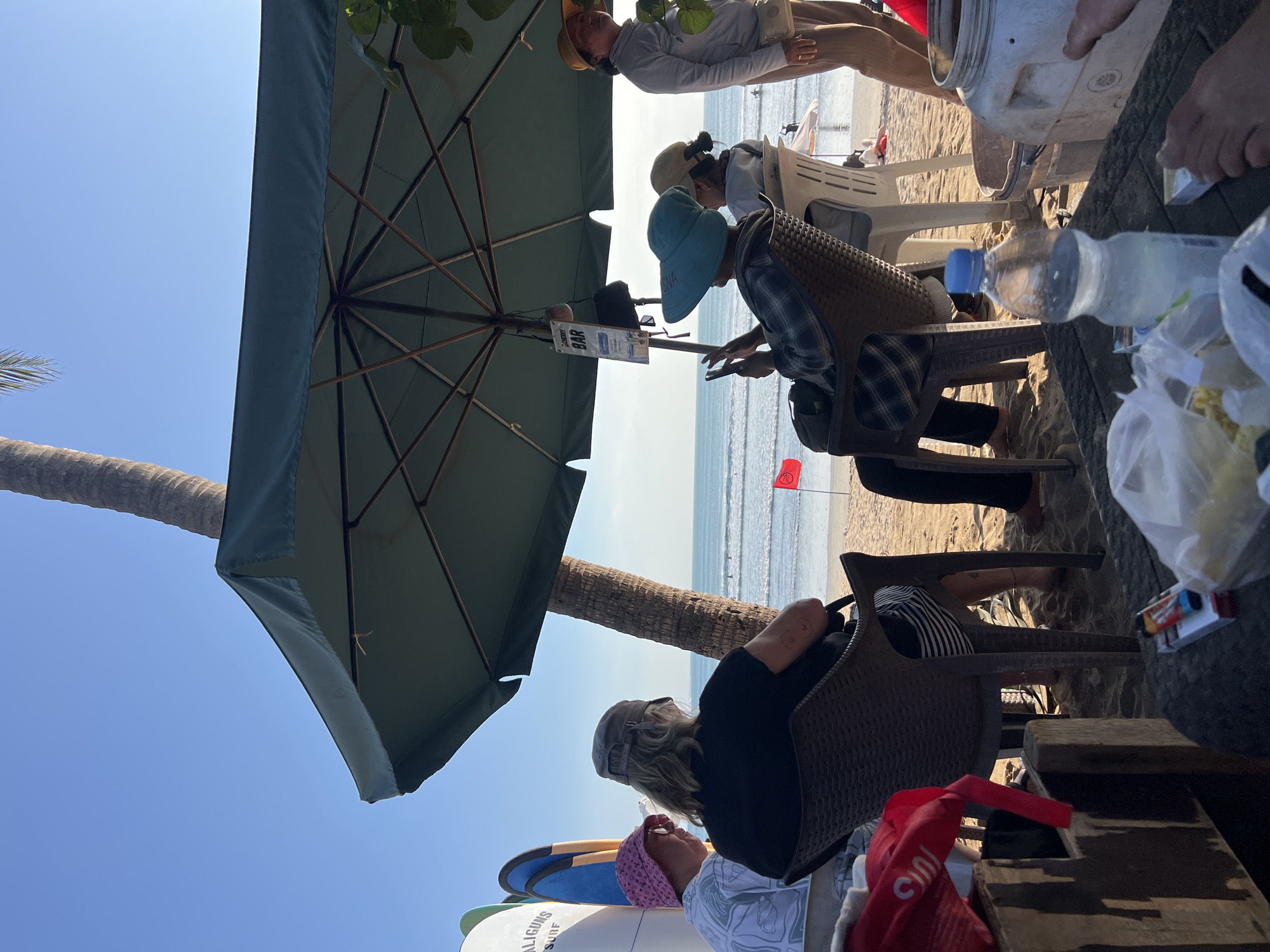 The image depicts a beachfront scene in Kuta, Indonesia, likely a cafe or bar area. Several individuals are seated under a large green umbrella, facing the ocean. In the foreground, a wooden table holds a partially consumed bottle of water, a pack of cigarettes, and some plastic-wrapped items. Two individuals are seated in chairs at the table, one wearing a pink bucket hat and the other a gray baseball cap. To the right of the table, a large white plastic container is visible. Further back, more people are seated in chairs, and some are standing. In the middle ground, the ocean stretches out with a few small waves and distant figures in the water. A red flag is planted in the sand. Surfing equipment, including surfboards, is leaning against a white structure on the left side of the image. A palm tree trunk is visible behind the umbrella. The sky is clear and blue.