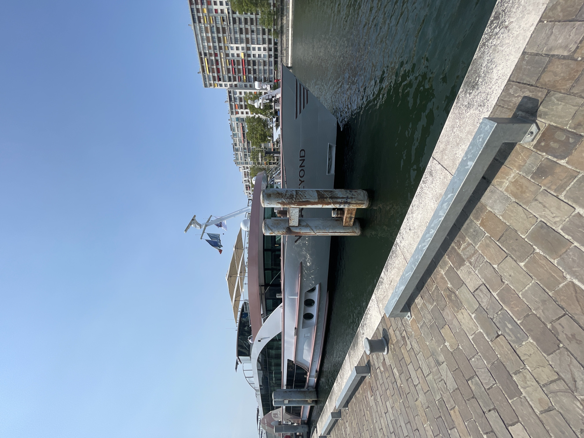 A luxury riverboat named "ELYON" is docked at a waterfront promenade in Paris, France. The boat is a light gray color with pink accents and has large glass windows. It is tied to a dock made of stone pavers, with metal bollards and a silver metal railing along the edge. In the background, several tall, modern apartment buildings with colorful awnings line the riverbank, and trees are visible. The sky is clear and blue, indicating a sunny day. There are no people visible in the frame.
