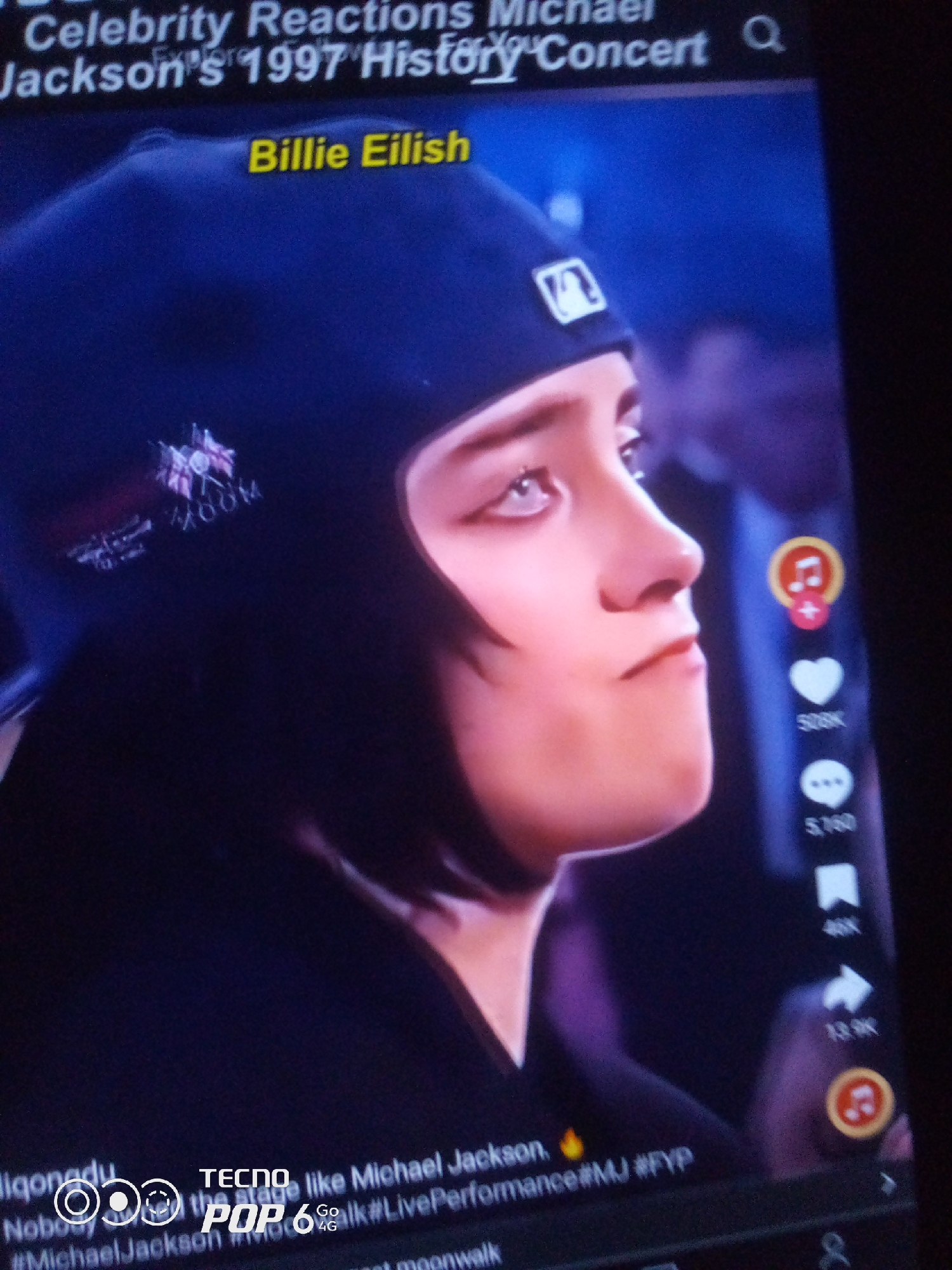 The image displays a screen, likely a smartphone or tablet, showing a video clip. The video features a person wearing a dark blue beanie with a white logo and a smaller embroidered patch. This person is identified as Billie Eilish by text overlaid on the screen. The text also indicates the video is about "Celebrity Reactions Michael Jackson's 1997 History Concert."
A timestamp and user handle are visible at the bottom of the screen: "liqonadu," "TECNO POP 6 Go 4G." Hashtags related to Michael Jackson and performance are also present. On the right side of the screen, there are social media interaction icons, including a music note with a red circle and plus sign, a heart with "508K" below it, a chat bubble with "5160" below it, and a bookmark icon with "46K" below it, indicating engagement metrics.
The image does not provide any information about the location being Howayi, Nigeria, or any other geographical details beyond what is presented on the screen itself. The scene is a digital display, not a physical location.
