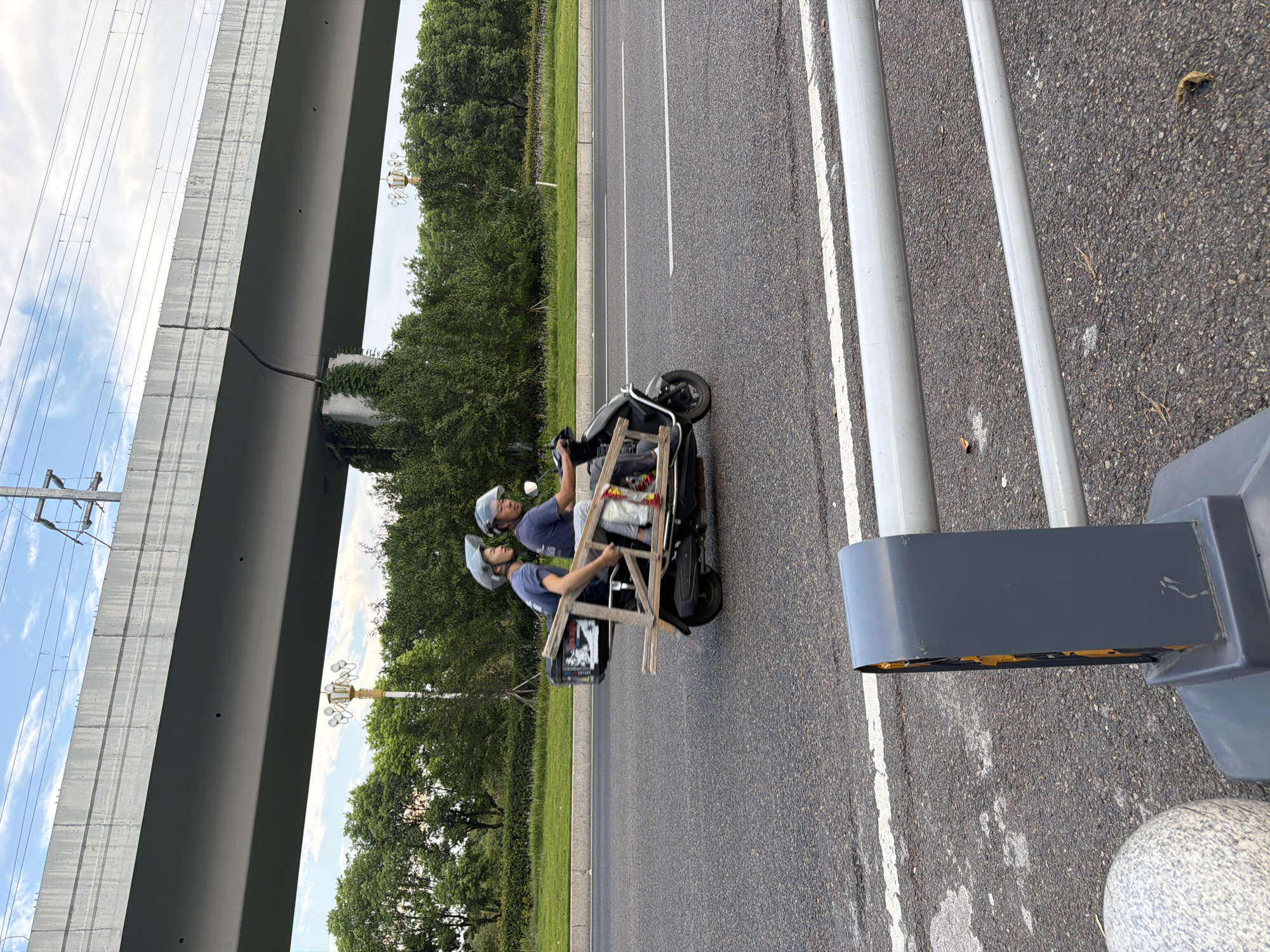 In Jiangjin, China, two individuals wearing helmets ride on a modified electric scooter on a paved road under an elevated concrete structure. The scooter appears to have been adapted to carry a ladder on its rear, supported by wooden beams. The driver is on the right, and a passenger is on the left. The road is marked with white lines. Trees and greenery line the roadside, with a cloudy sky overhead. In the foreground, a grey and white striped bollard and a metal barrier are visible.